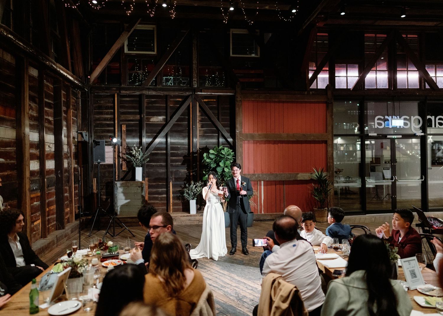 A group gathers around a long rustic wooden table in a barn-like setting for a communal dining experience.