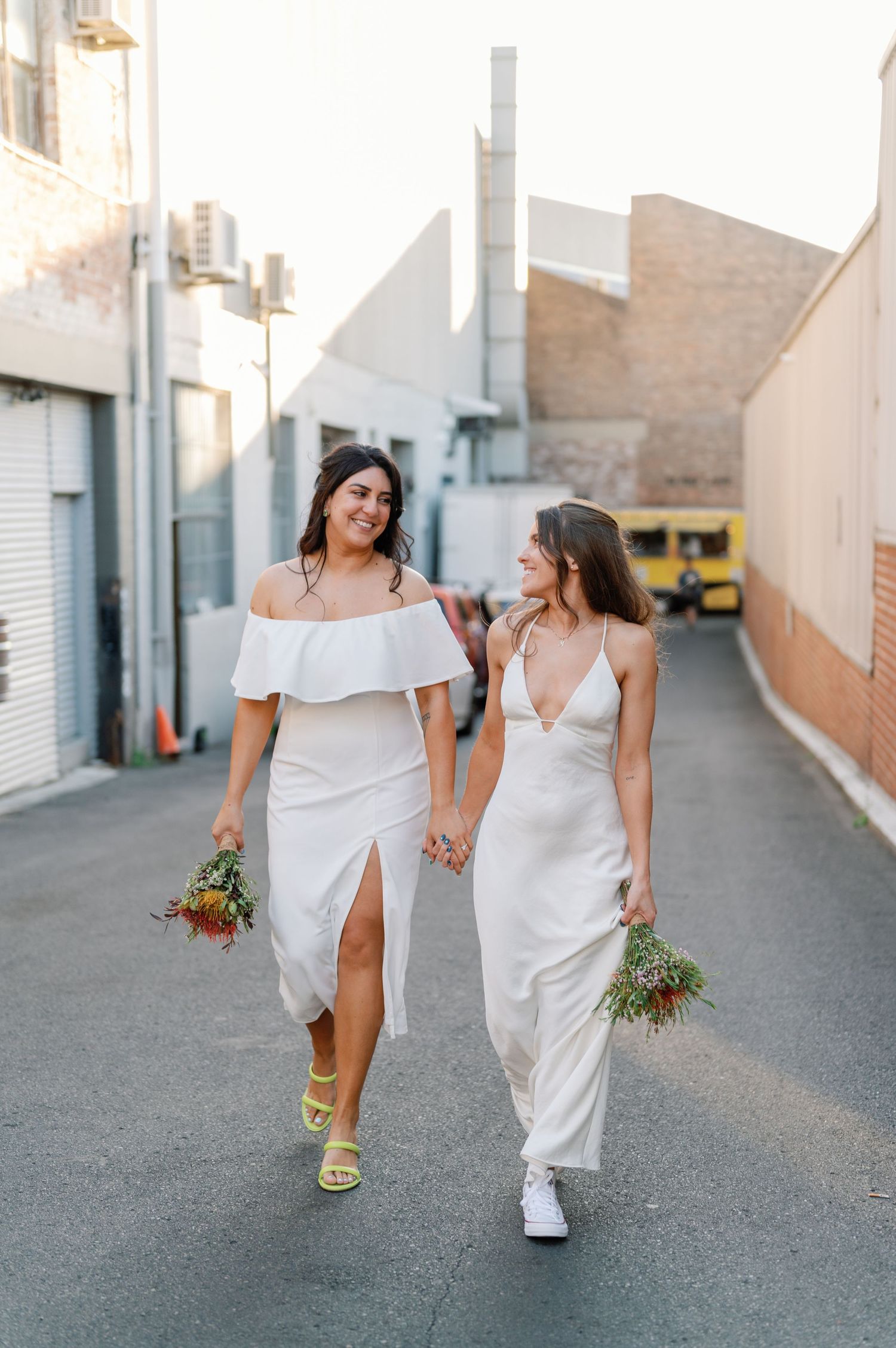 Two people in white dresses walk down an urban alley holding hands and bouquets while wearing contrasting footwear.