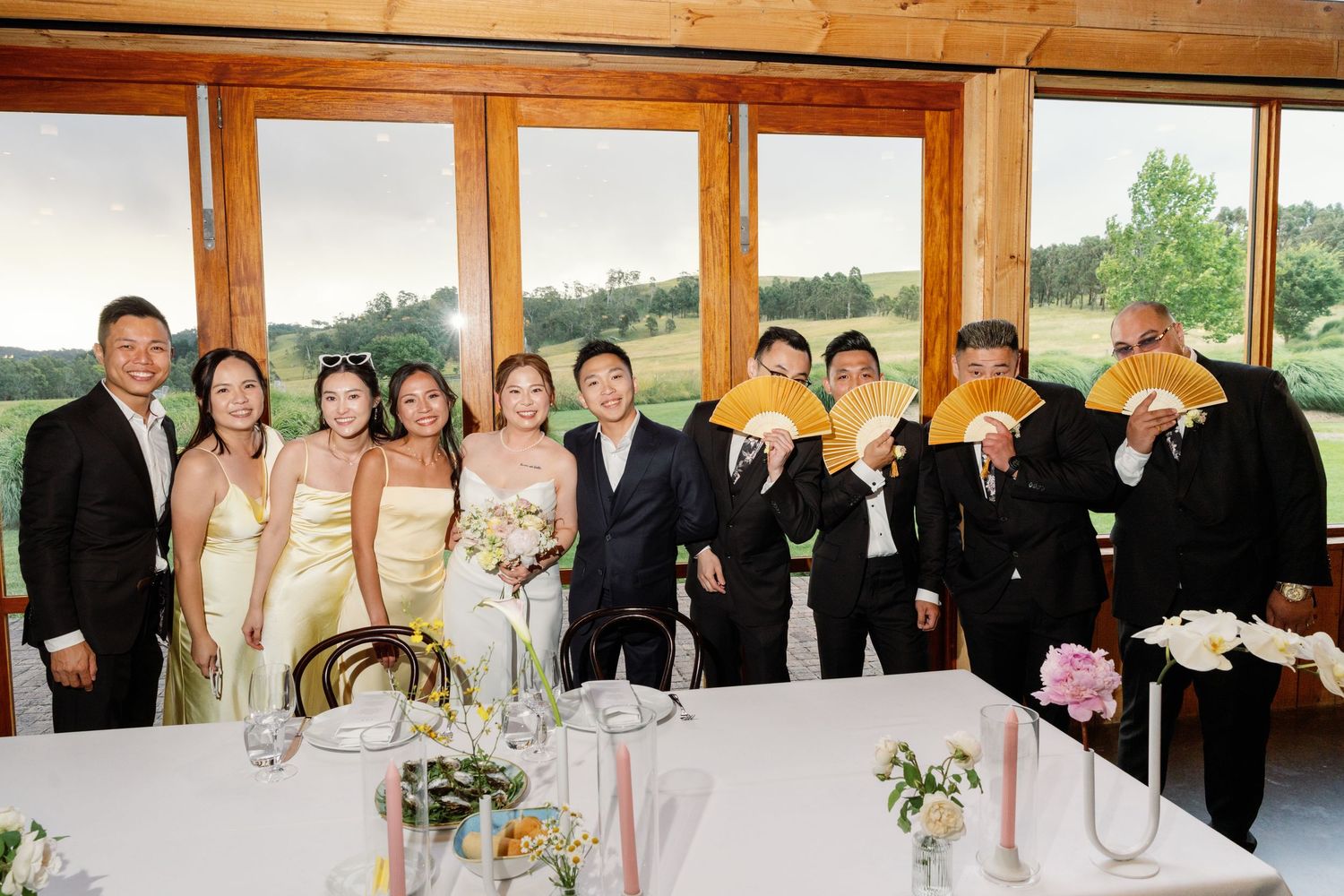 A panoramic wedding party group photo with bridesmaids and groomsmen lined up in a rustic wooden venue.