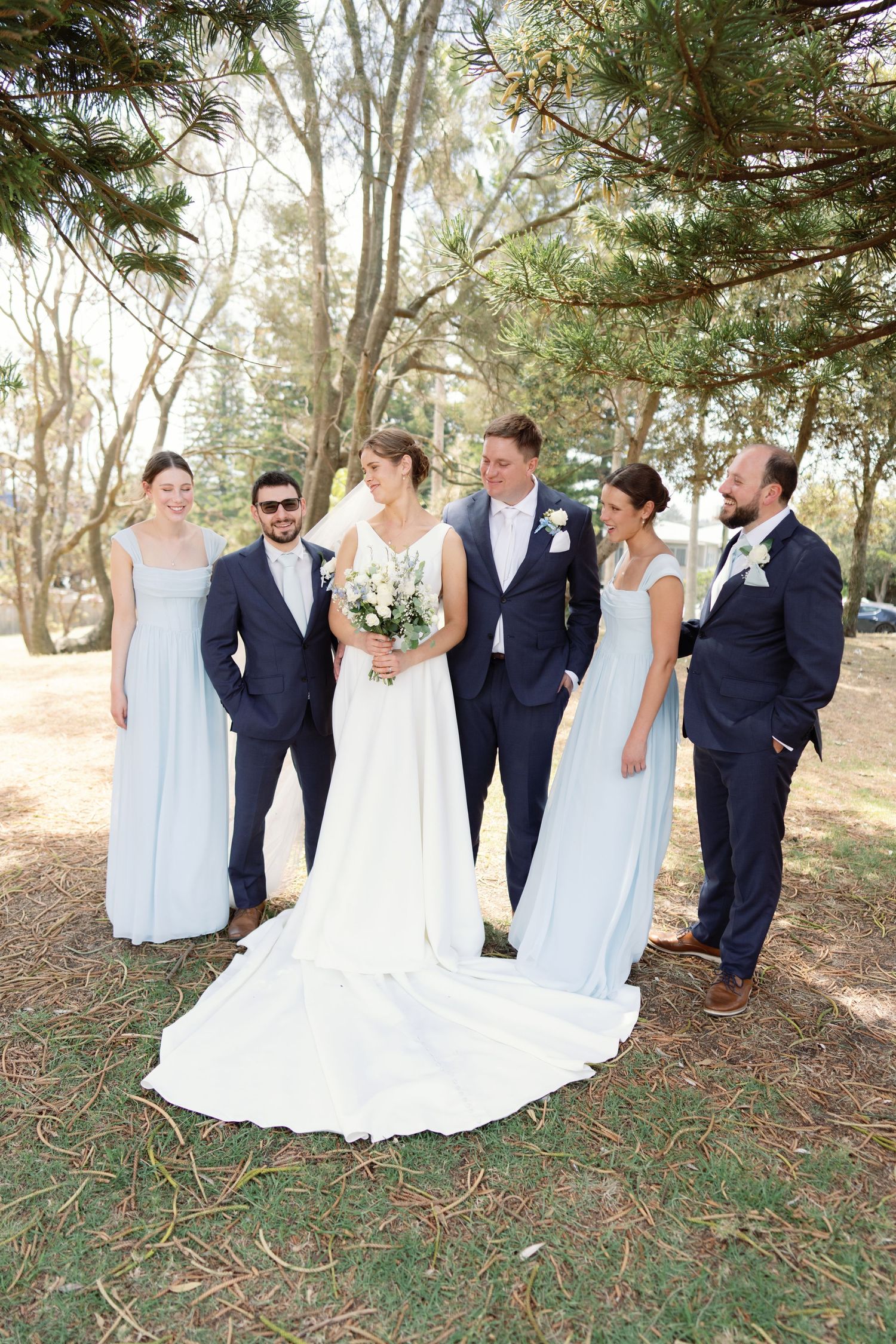 Wedding party in navy suits and light blue dresses poses together outdoors among trees.