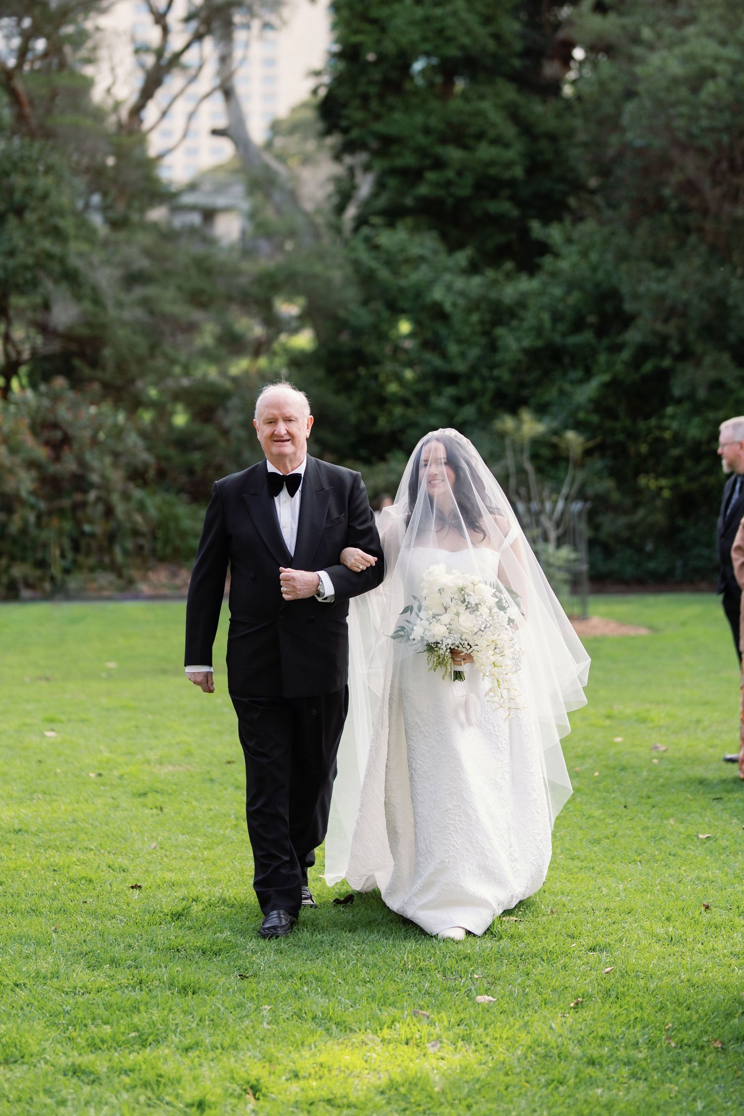 Father walks bride down outdoor aisle in white gown and veil, holding bouquet on green lawn.