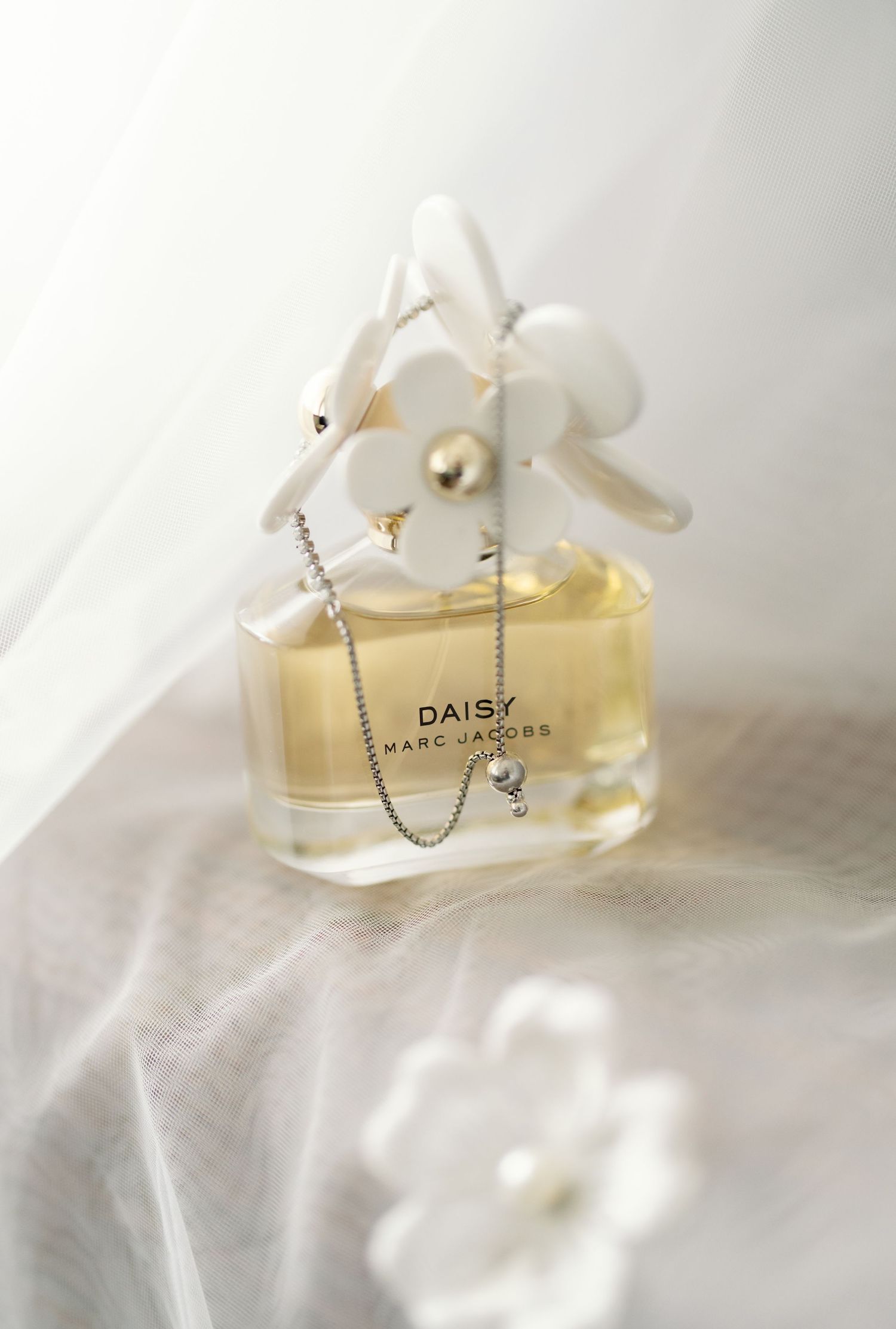 Elegant perfume bottle labeled 'Daisy' sits on white fabric with delicate silver chain details.