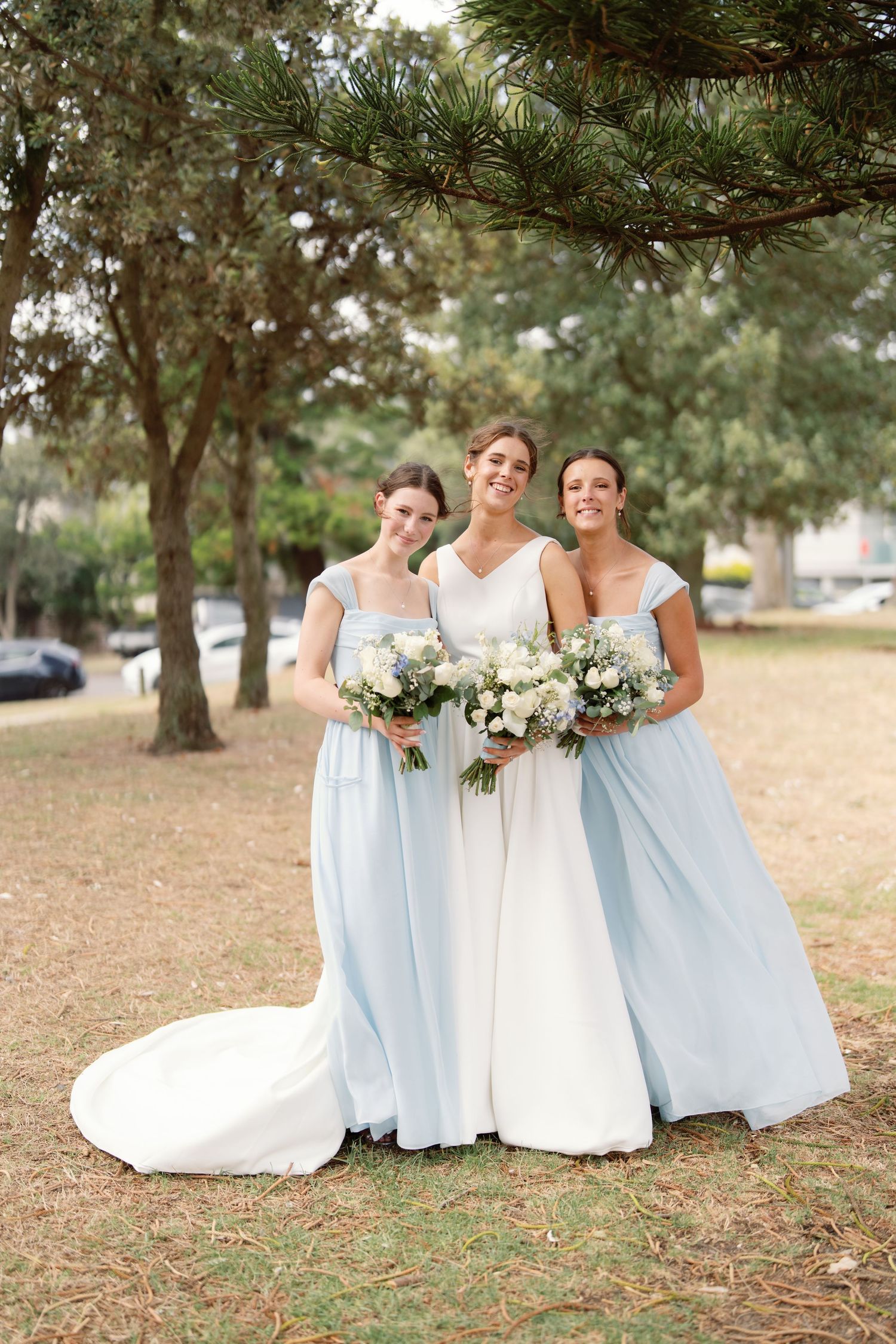 A bridal party in light blue bridesmaid dresses and white wedding gown pose together outdoors under pine trees.
