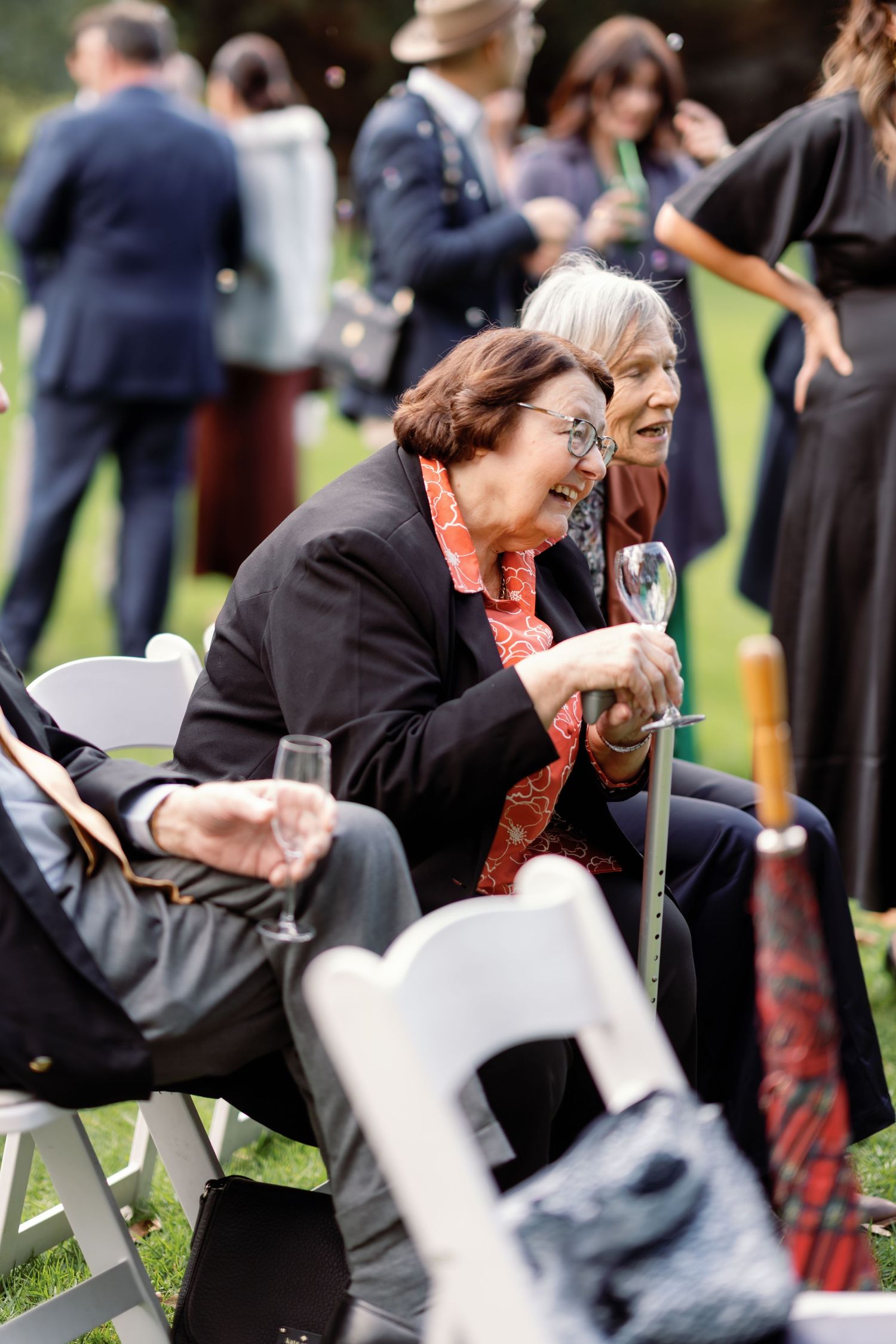 Two elderly women laughing and enjoying champagne at an outdoor wedding reception seated on white chairs.
