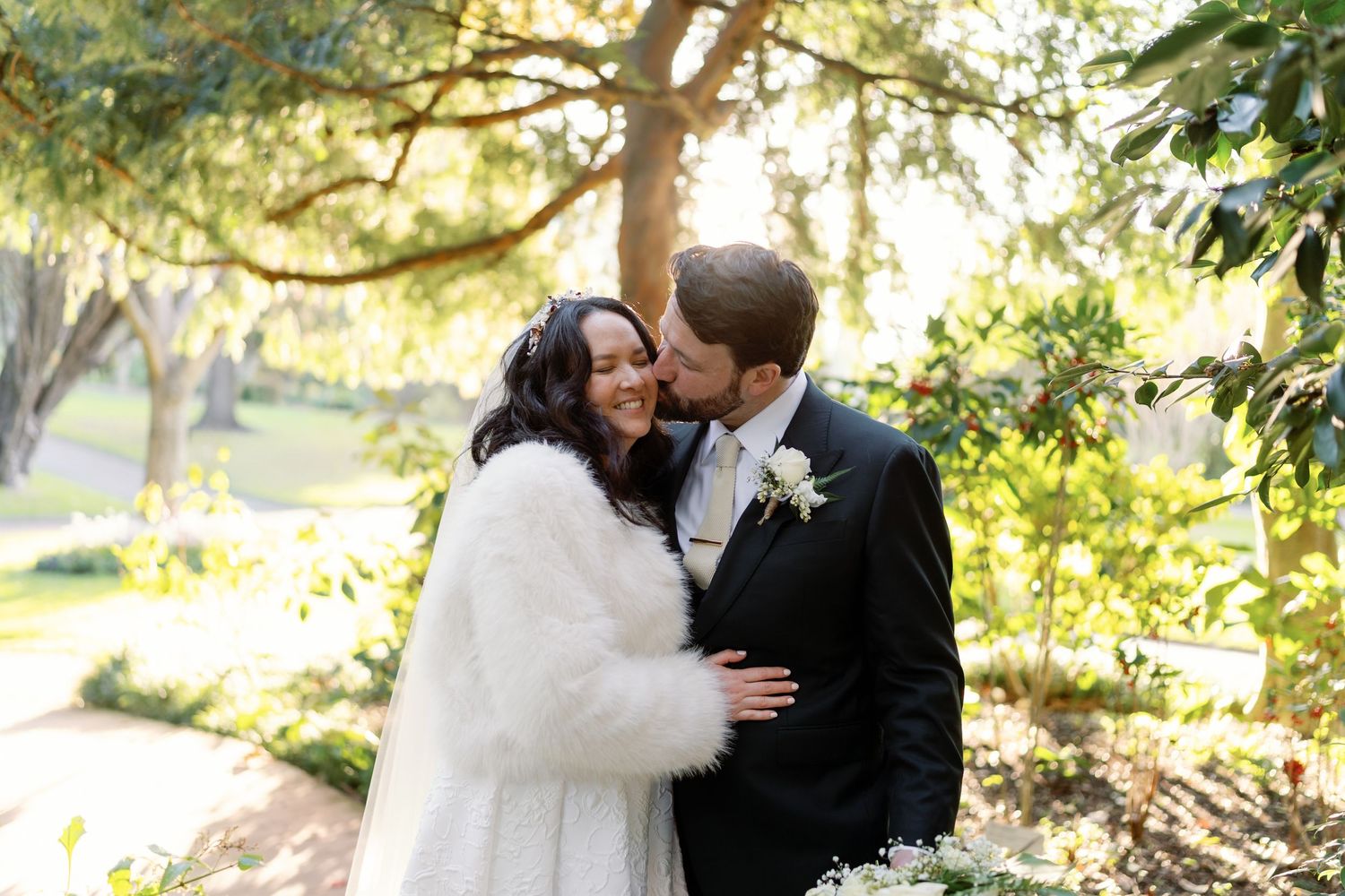 Groom tenderly kisses bride wearing white fur wrap in sunlit garden wedding portrait.