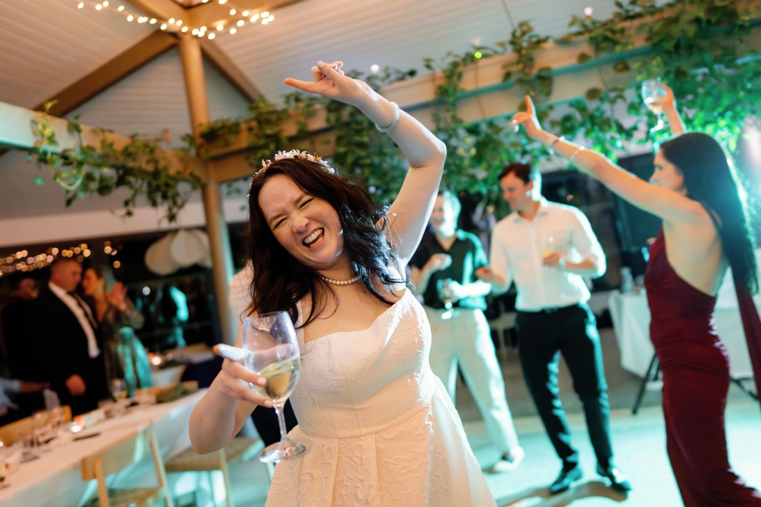 Joyful bride in white gown dances with raised arm holding wine glass at reception.