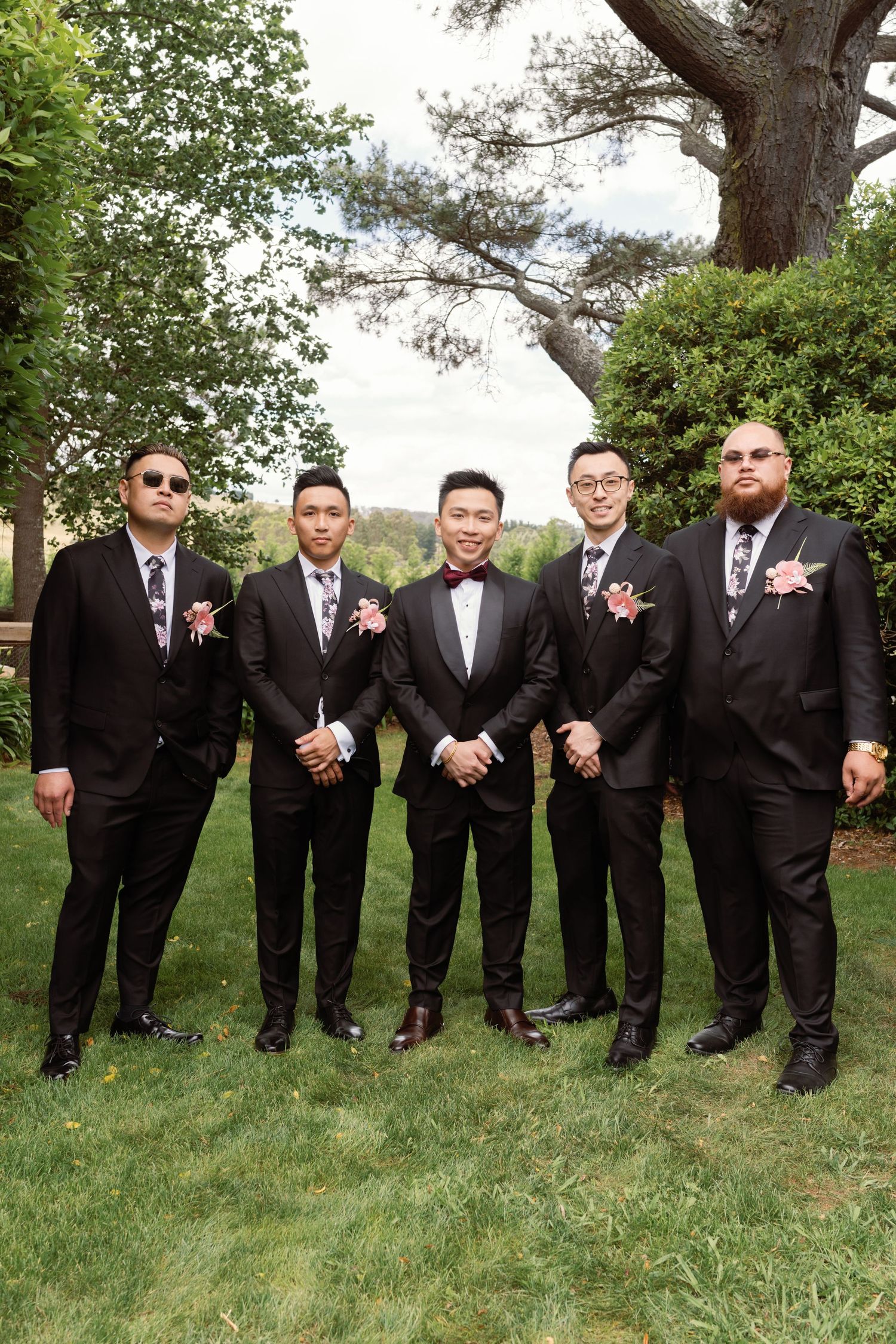 Groomsmen in black tuxedos pose playfully on grass under trees.
