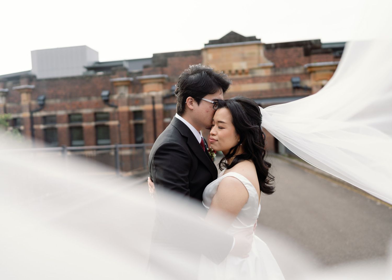 A romantic moment captured against brick building backdrop with flowing white veil.