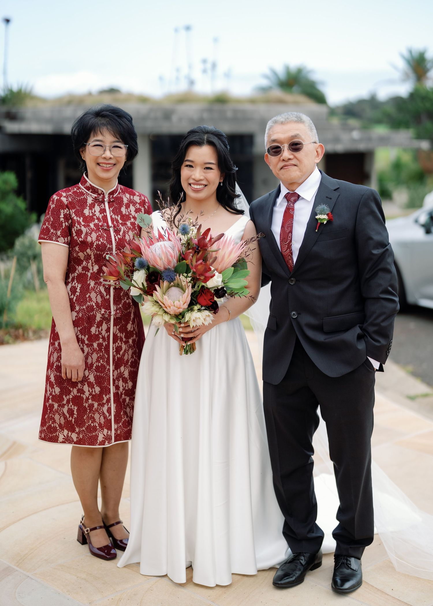 A bride in a white dress holds a red and pink bouquet while posing with family members at an outdoor wedding.