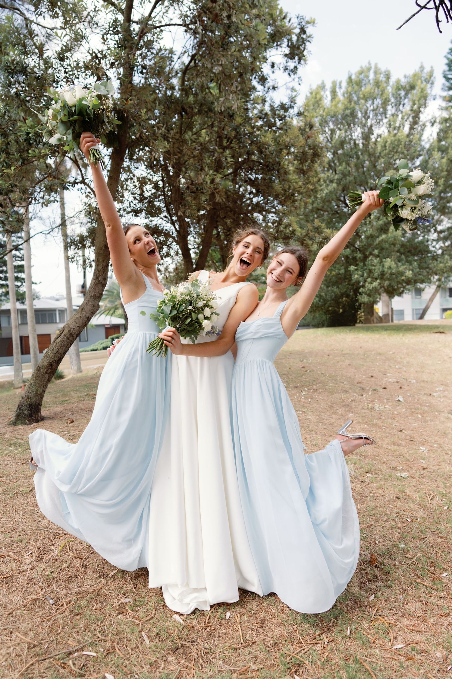 Bridal party in white and light blue dresses posing joyfully on grassy hillside.