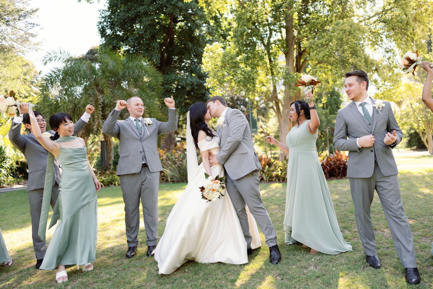 Wedding party in light gray and sage green attire poses playfully in a sunlit garden setting.