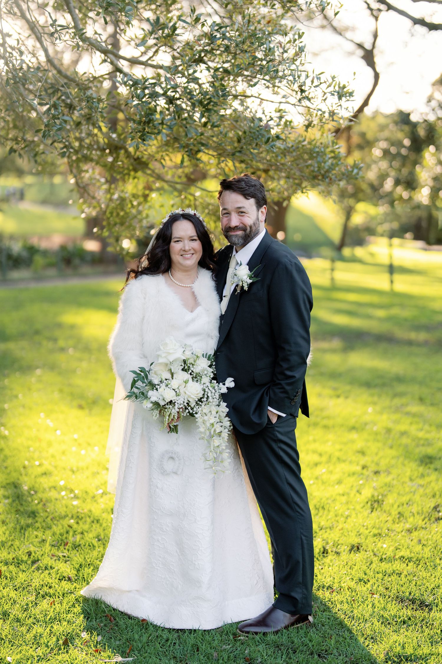 Bride and groom smiling together outdoors on green grass under olive trees in wedding attire with white bouquet.