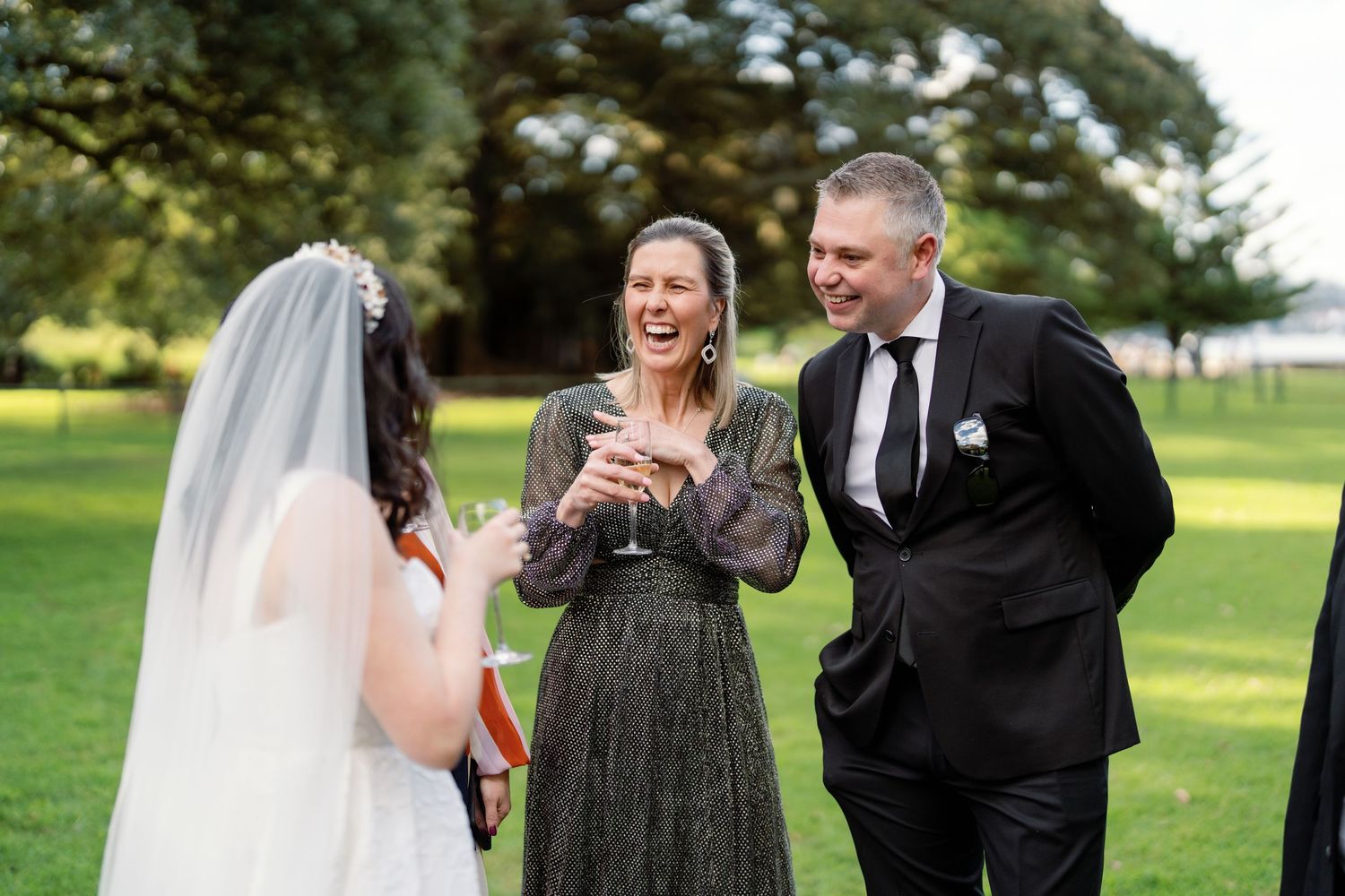 A bride in a veil laughs with two guests in formal attire on a green lawn at a wedding reception.