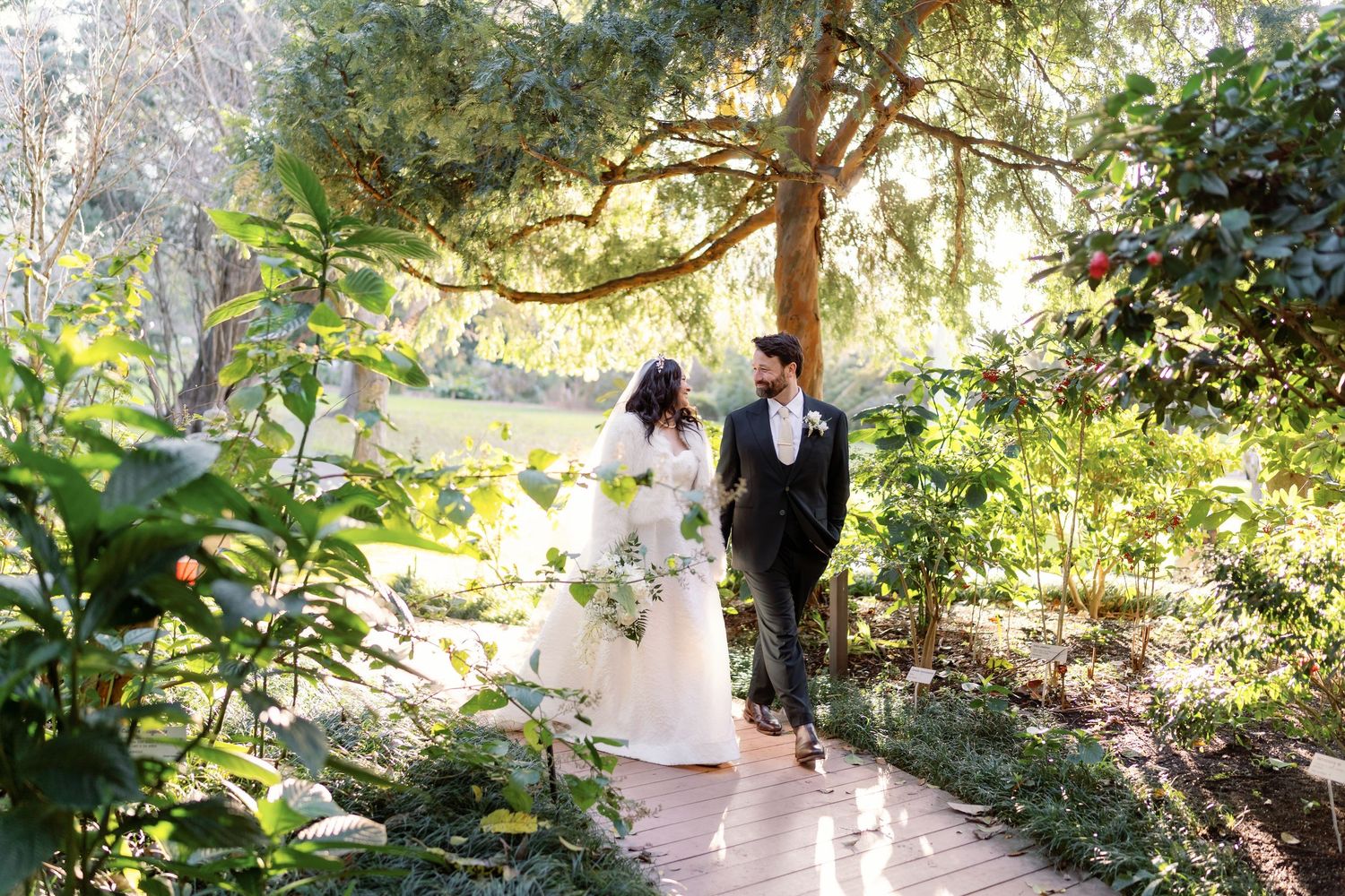 Bride and groom walk hand in hand along wooden path through lush green garden.