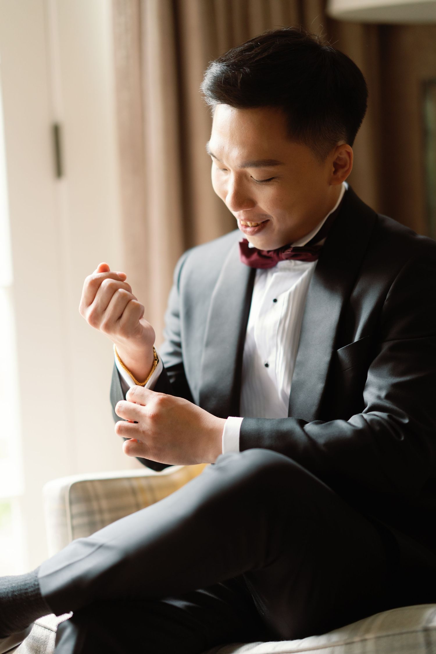 A person in a formal tuxedo and bow tie smiles while looking at their phone in a warmly lit room.