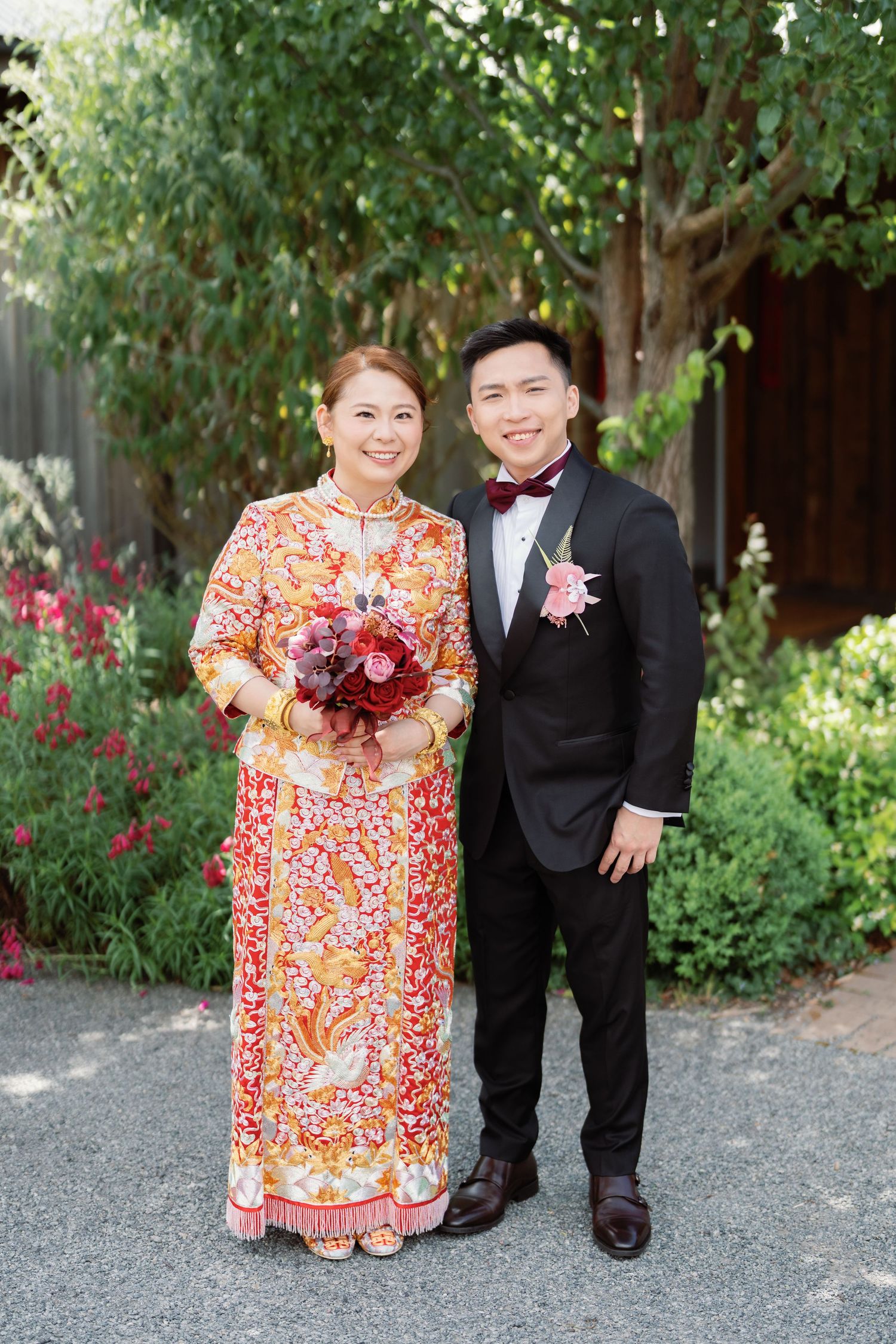 A couple poses outdoors with a burgundy bouquet against a garden backdrop.