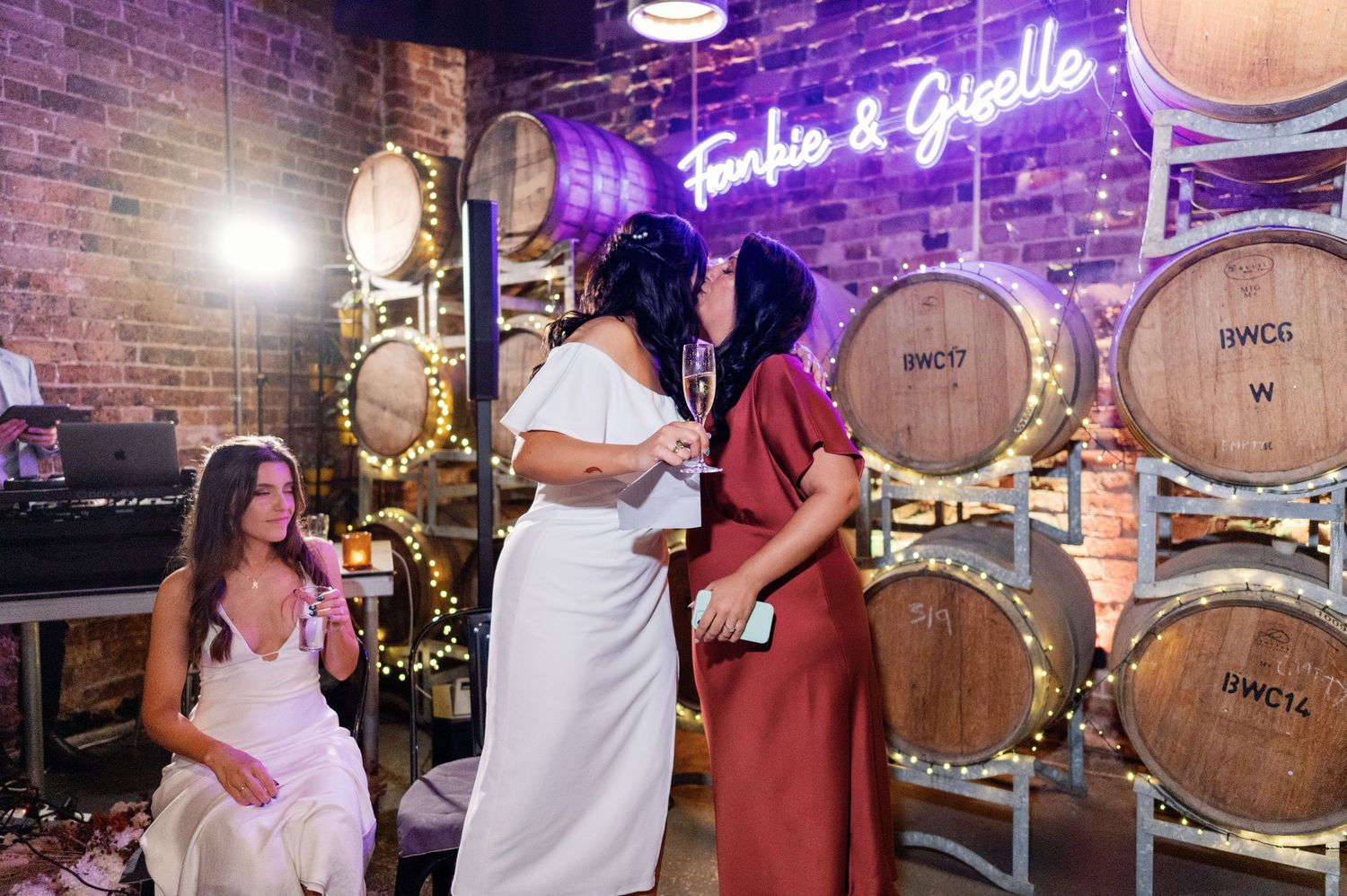 A group in white dresses poses in front of wooden wine barrels under purple neon lighting.