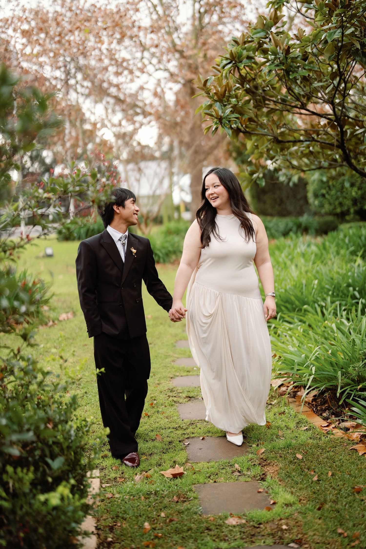 A couple in wedding attire walks hand-in-hand through a lush garden path, smiling at each other.