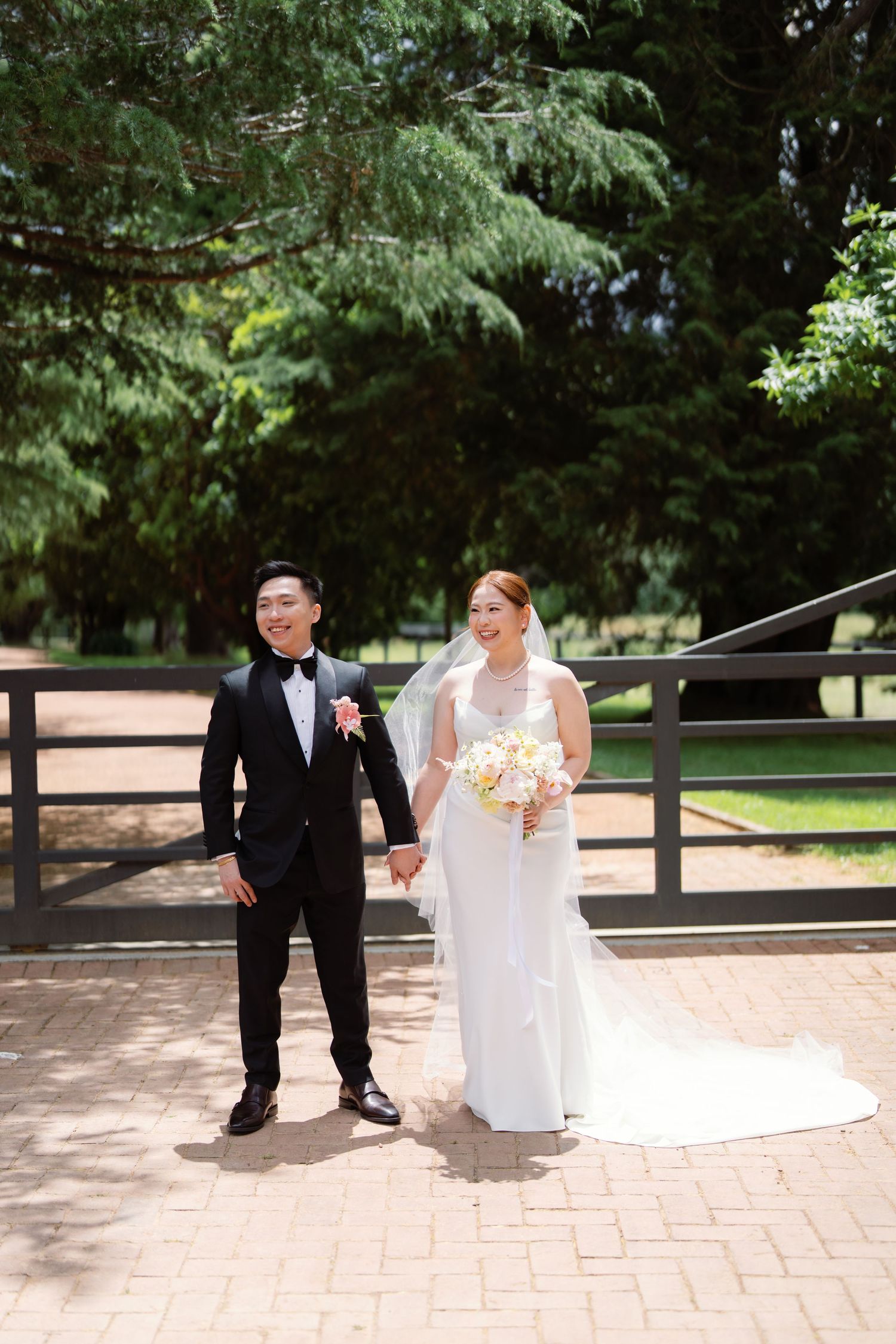 Wedding couple sits playfully on brick pavement surrounded by evergreen trees.