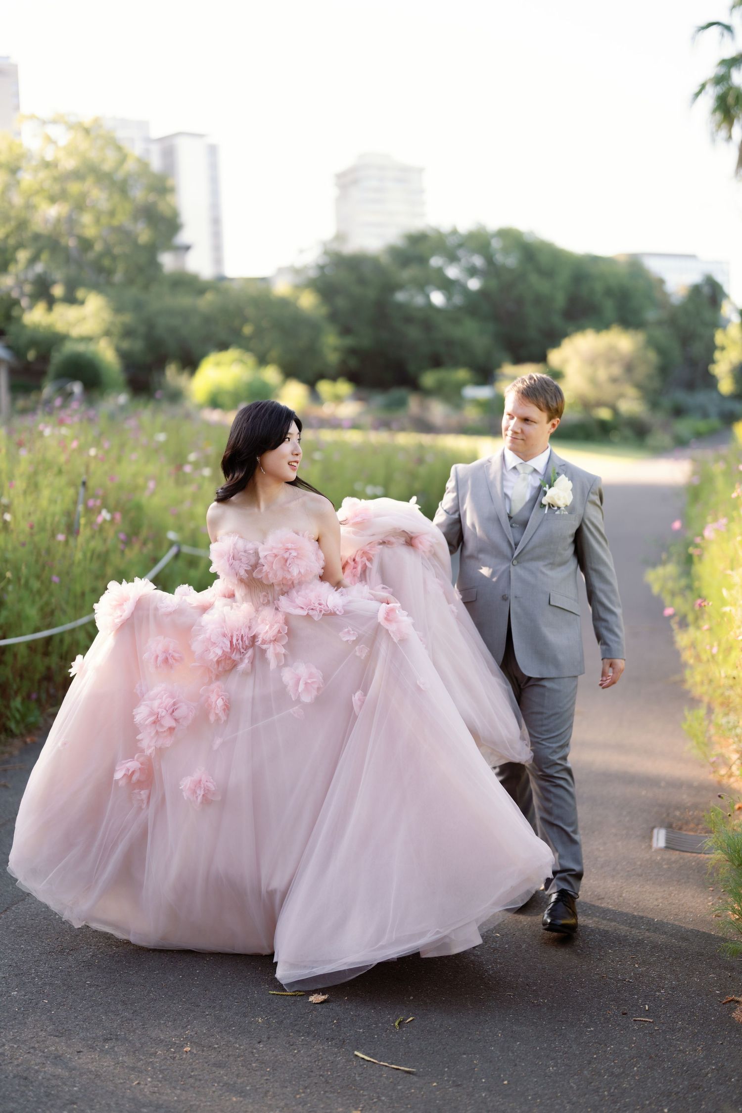 Couple in grey suit and pink floral wedding gown dancing on path among wildflowers.