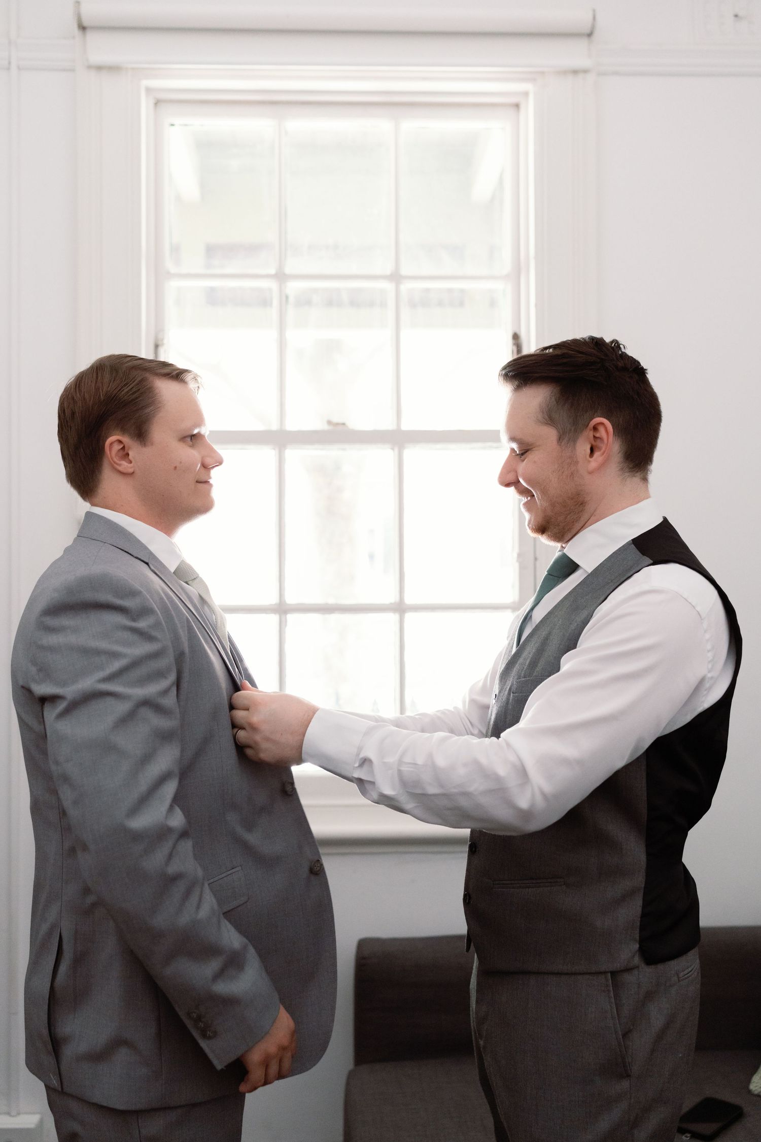 Groomsmen help each other adjust their ties and suits in a bright indoor setting before a wedding ceremony.