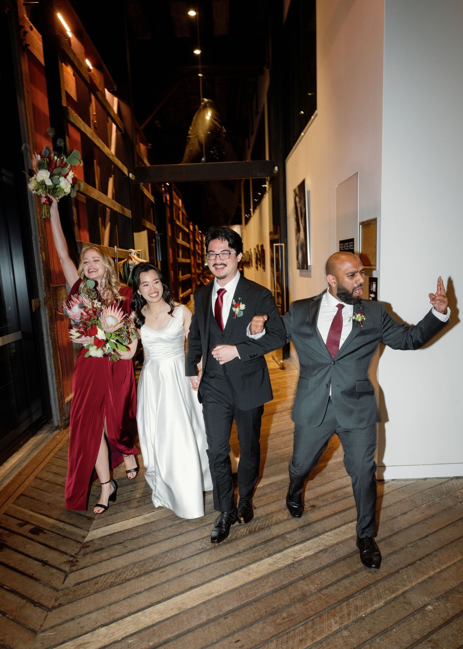 Wedding party walks and poses energetically down a wooden hallway with dark walls.