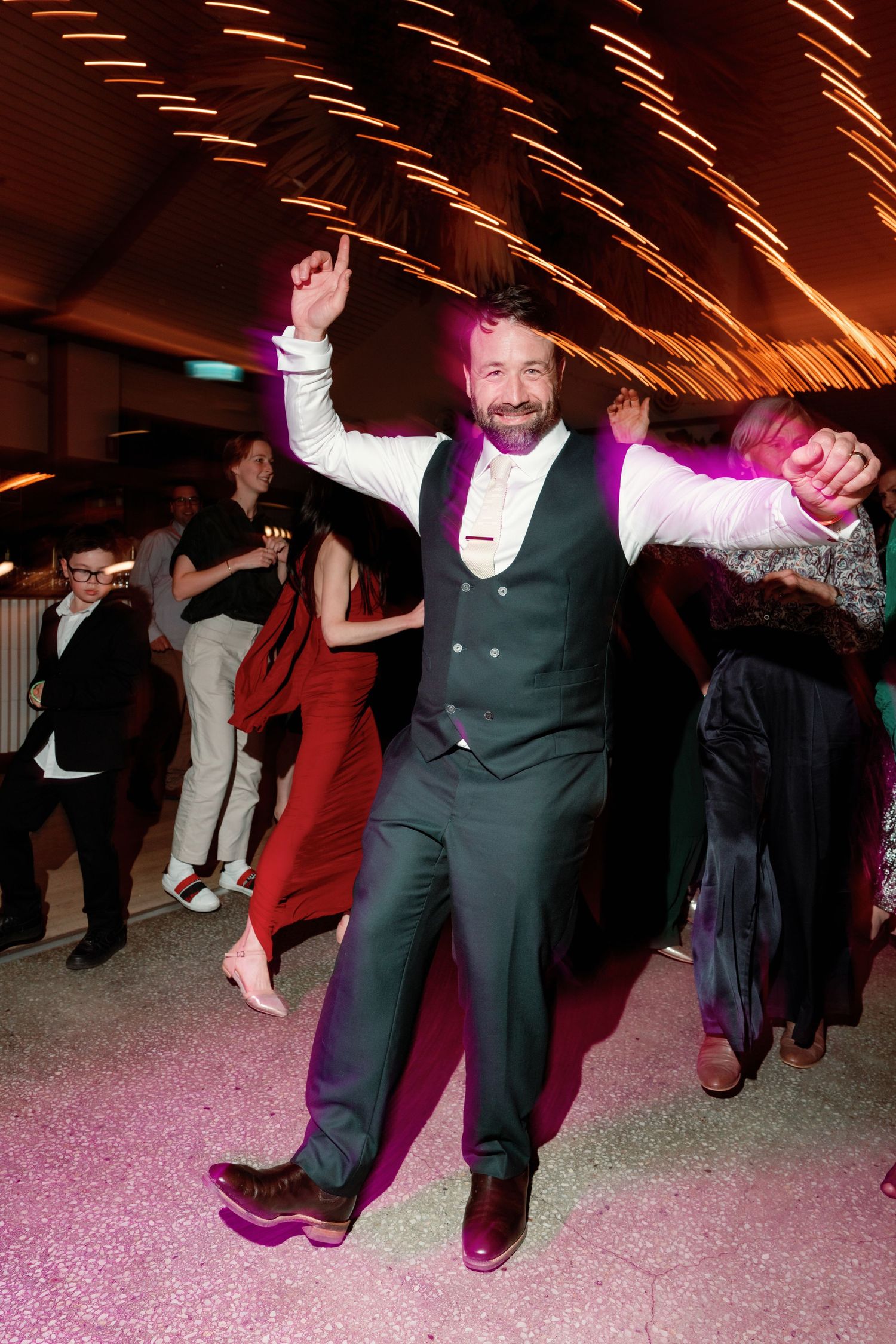 Groom in grey vest and trousers dancing energetically on lit dance floor at wedding reception.