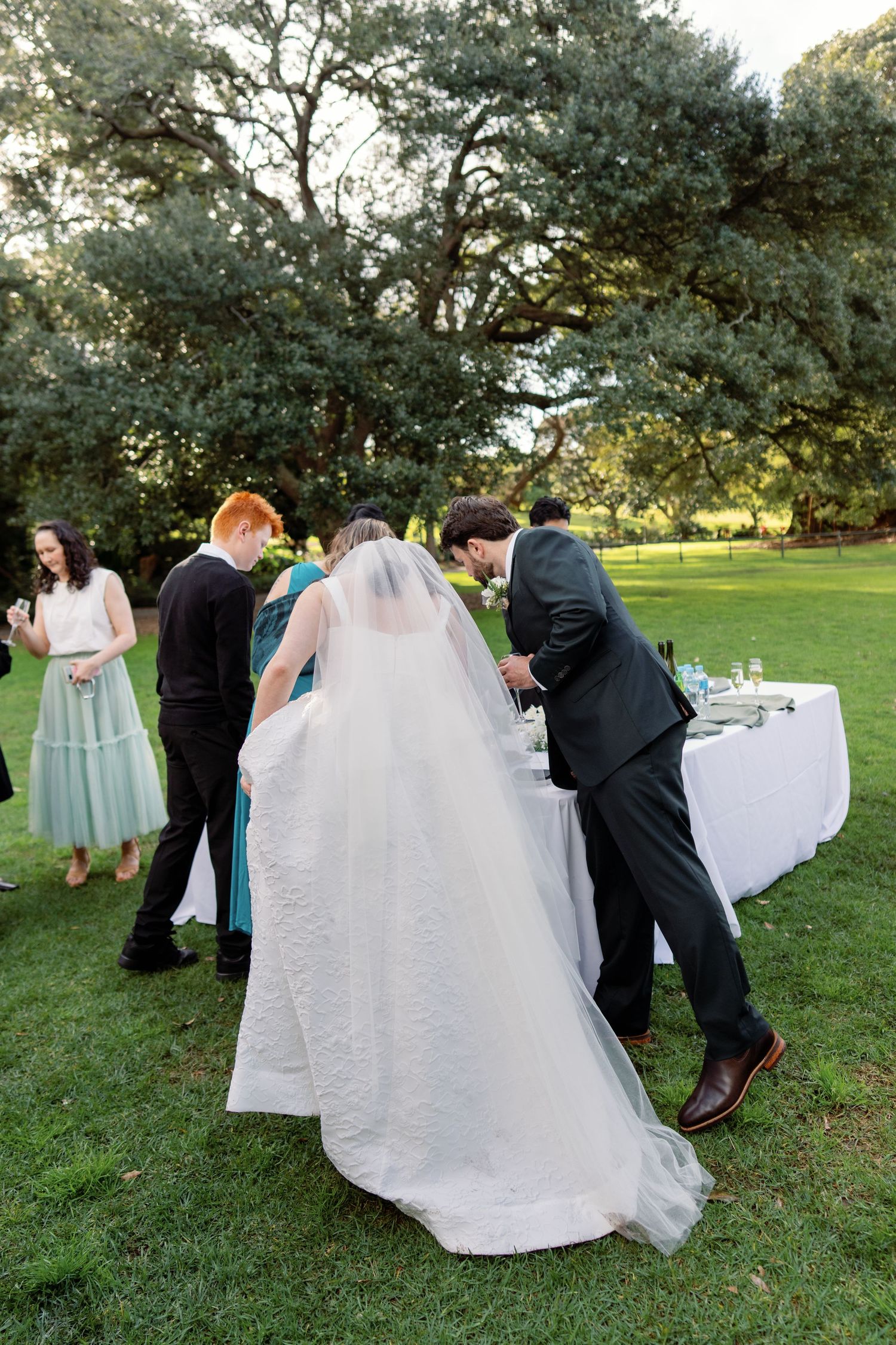 Bride in white gown and veil stands with groom at outdoor wedding reception table on lush green lawn.