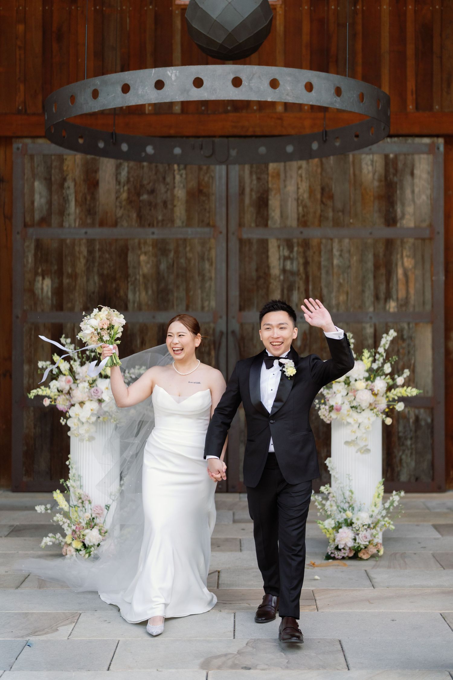 Newlyweds celebrate with joyful poses inside rustic barn decorated with white floral arrangements.