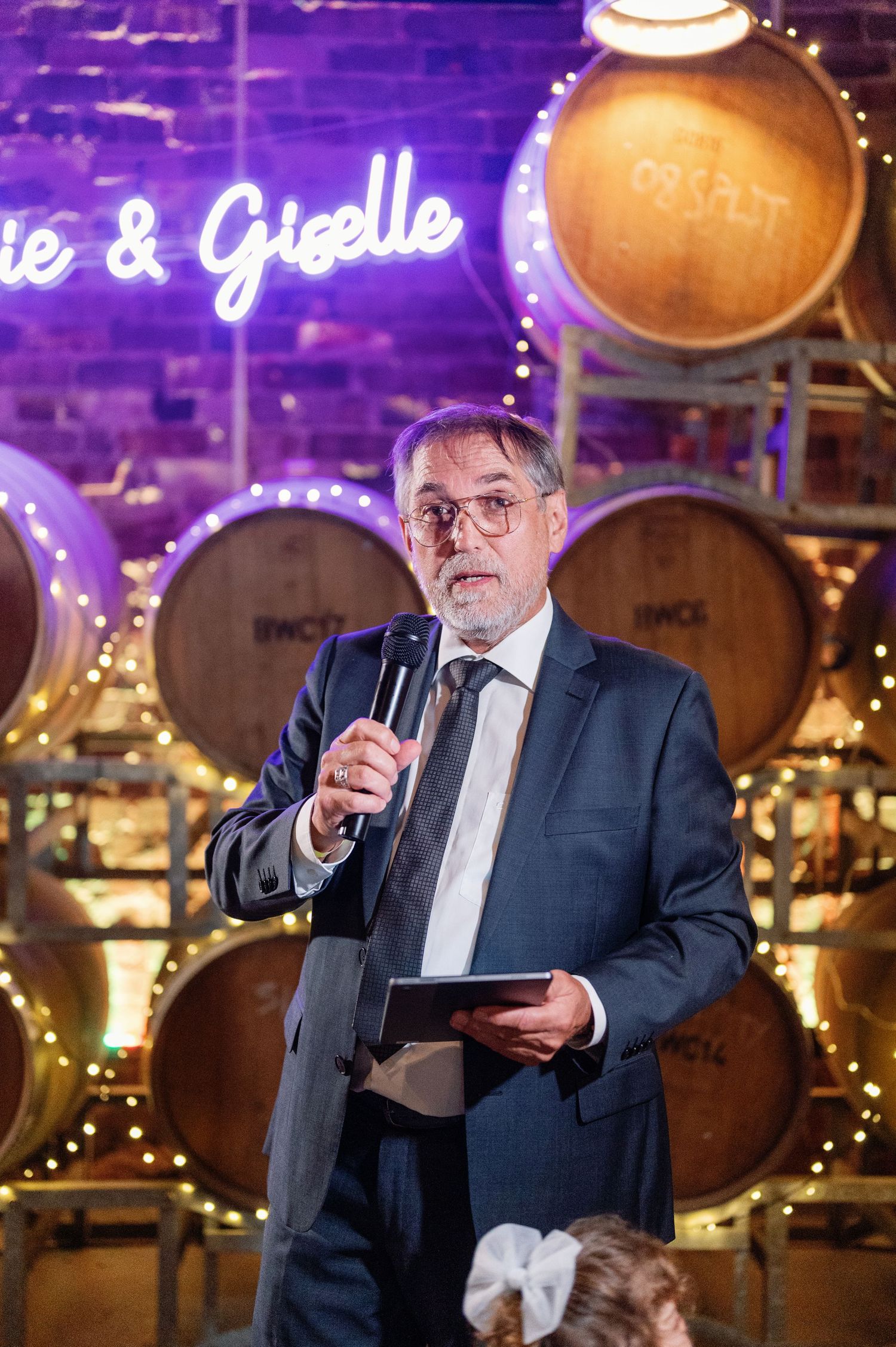 Speaker giving a toast in formal attire against purple-lit barrel room backdrop.