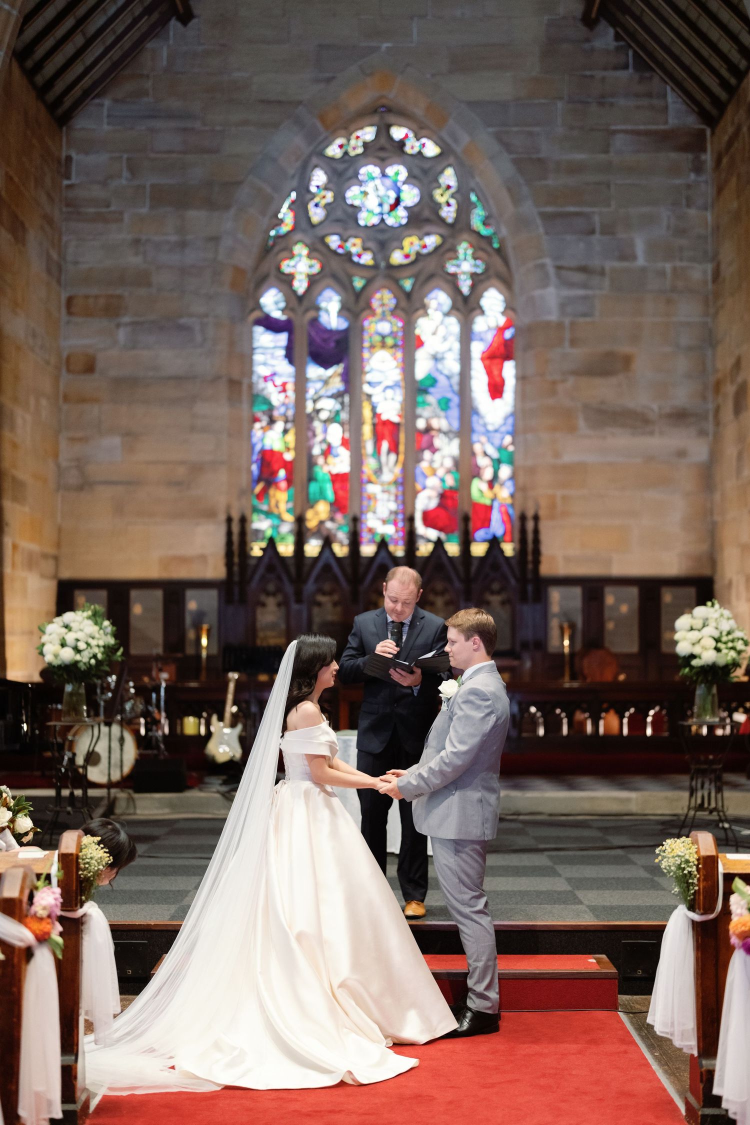 A wedding ceremony takes place in a church with a beautiful stained glass window and red carpet aisle.