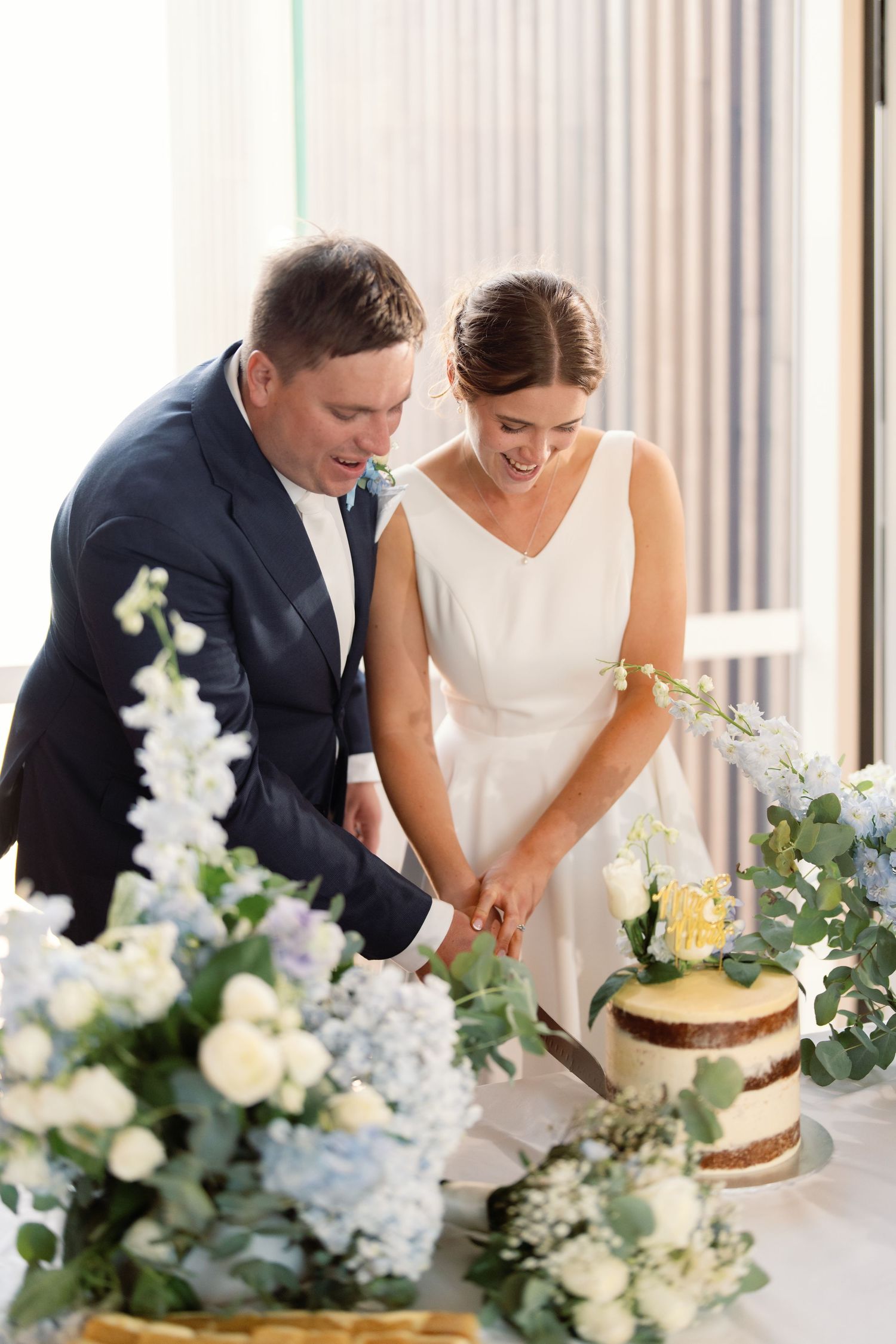Couple cutting their rustic wedding cake surrounded by white floral arrangements and greenery.