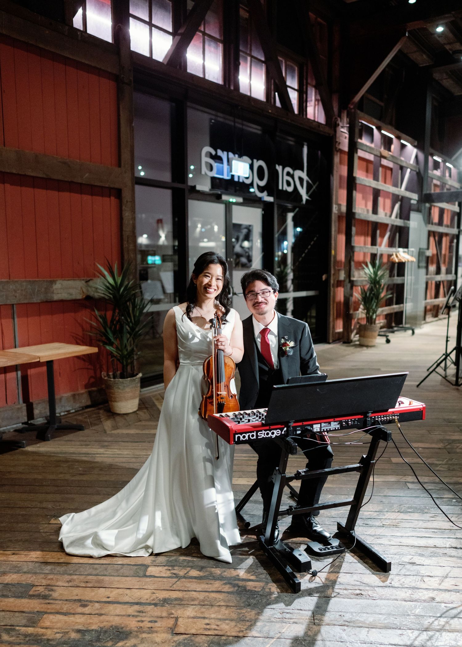 A couple poses near a keyboard piano on a wooden deck with red walls.