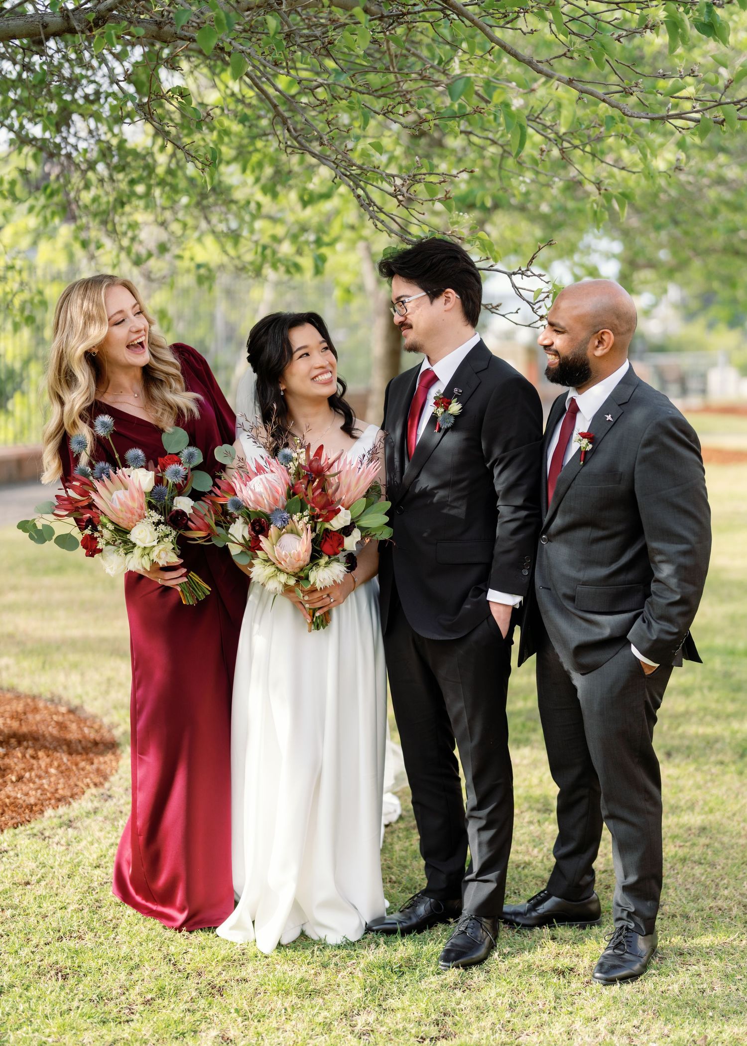Wedding party in burgundy dresses and black suits laughing together during outdoor photo session.