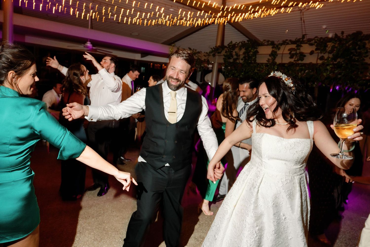 Bride in white gown and groom in vest dance joyfully with guests at elegant wedding reception venue.