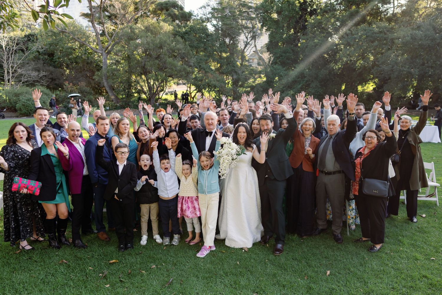 Large wedding group photo outdoors on green lawn with bride and groom surrounded by cheering guests waving hands.