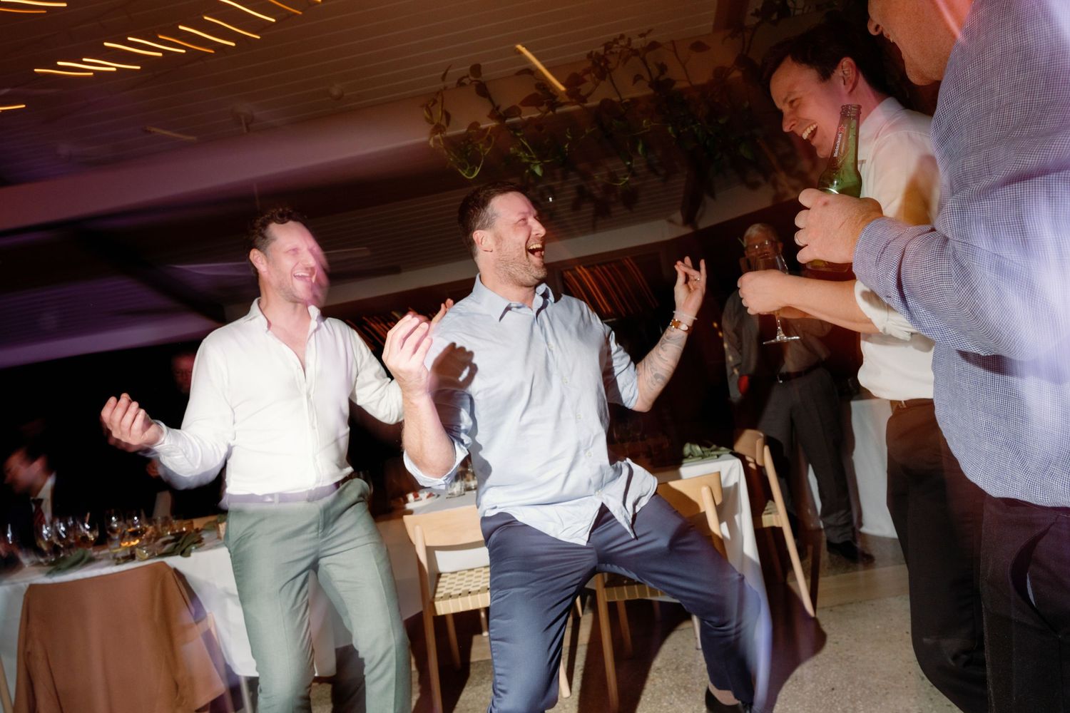 Men laughing and dancing joyfully at an indoor celebration or wedding reception party.