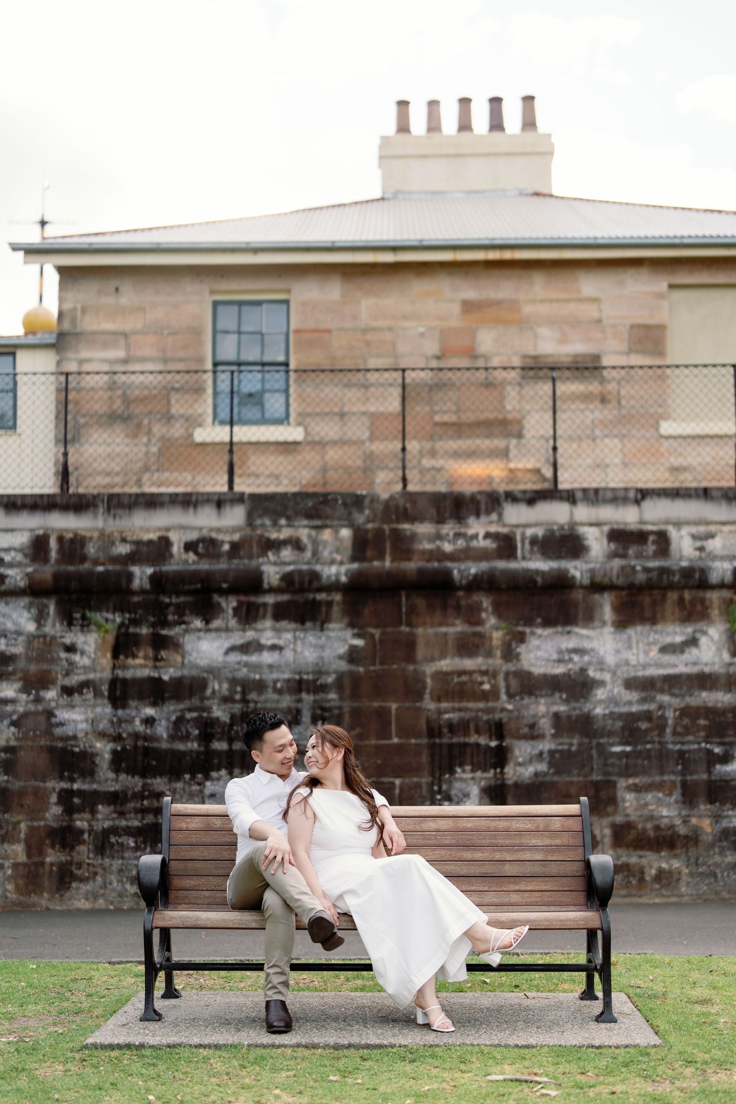 A couple in wedding attire share romantic moments while sitting on a park bench in front of a stone wall.