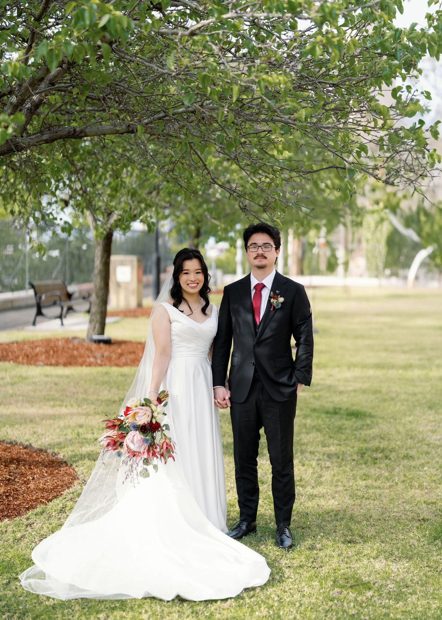 A bride in white gown and veil walks with companion on grassy lawn with trees.