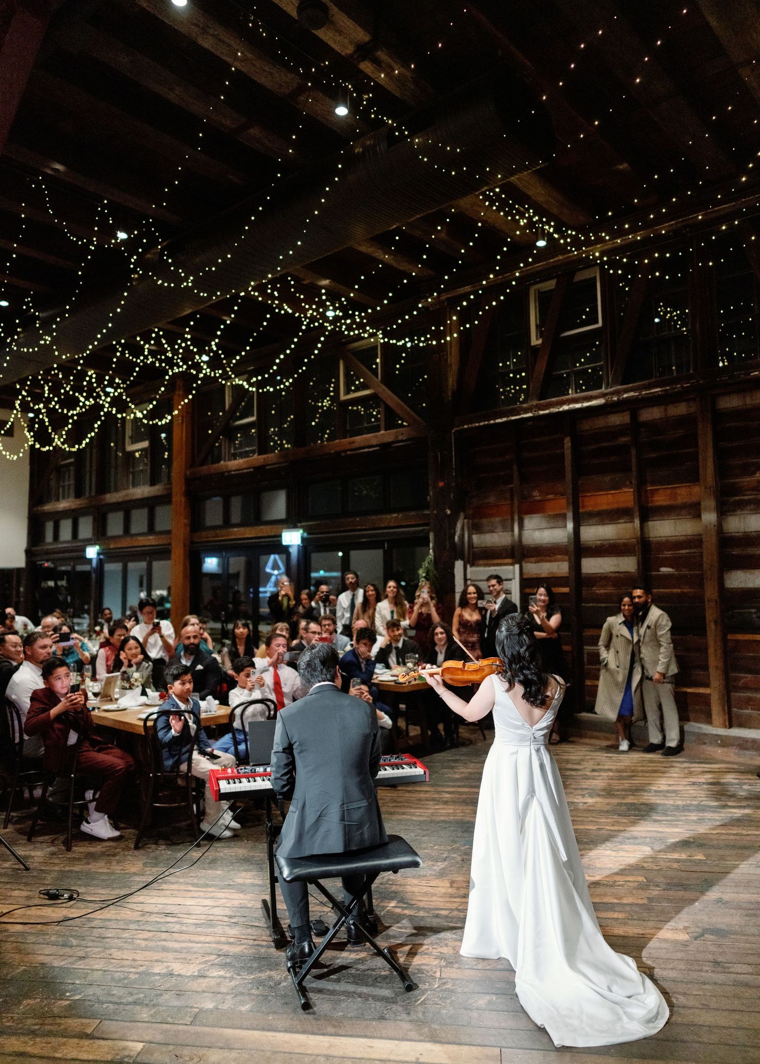 Panoramic view of a rustic barn wedding reception with string lights and couples dancing on wooden floor.