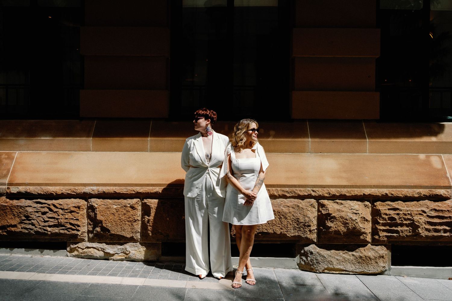 A couple poses against a stone building wall in white wedding attire on a sunny day.