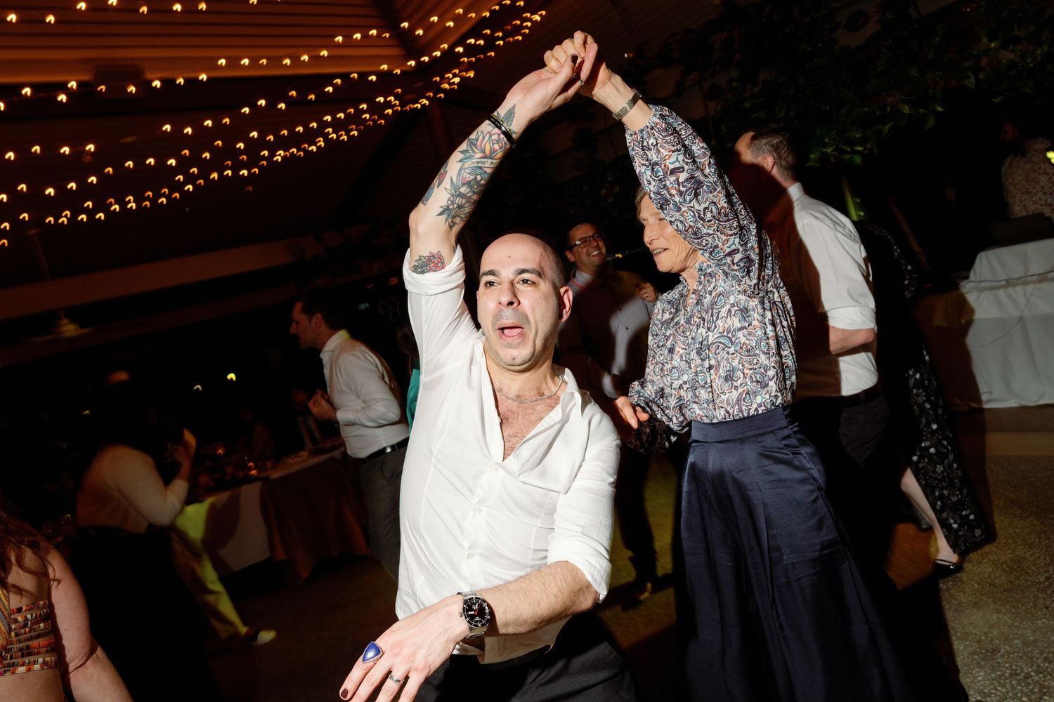Man with tattoos dancing energetically with a woman at an outdoor wedding reception under string lights.