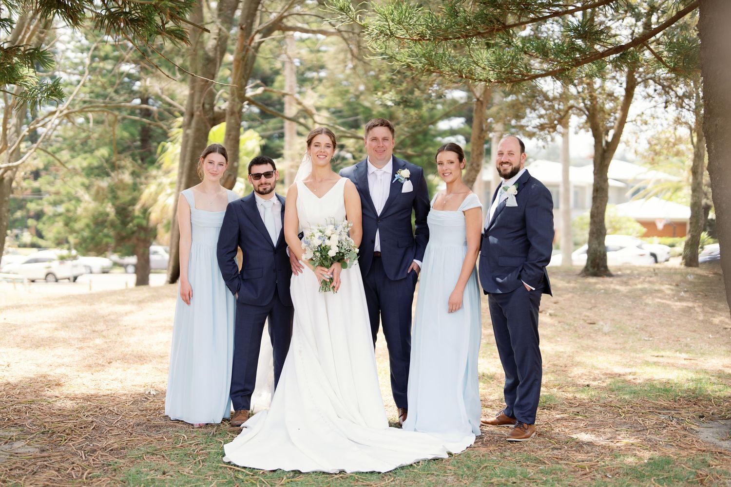 Wedding party poses for photos outdoors under trees in formal attire.