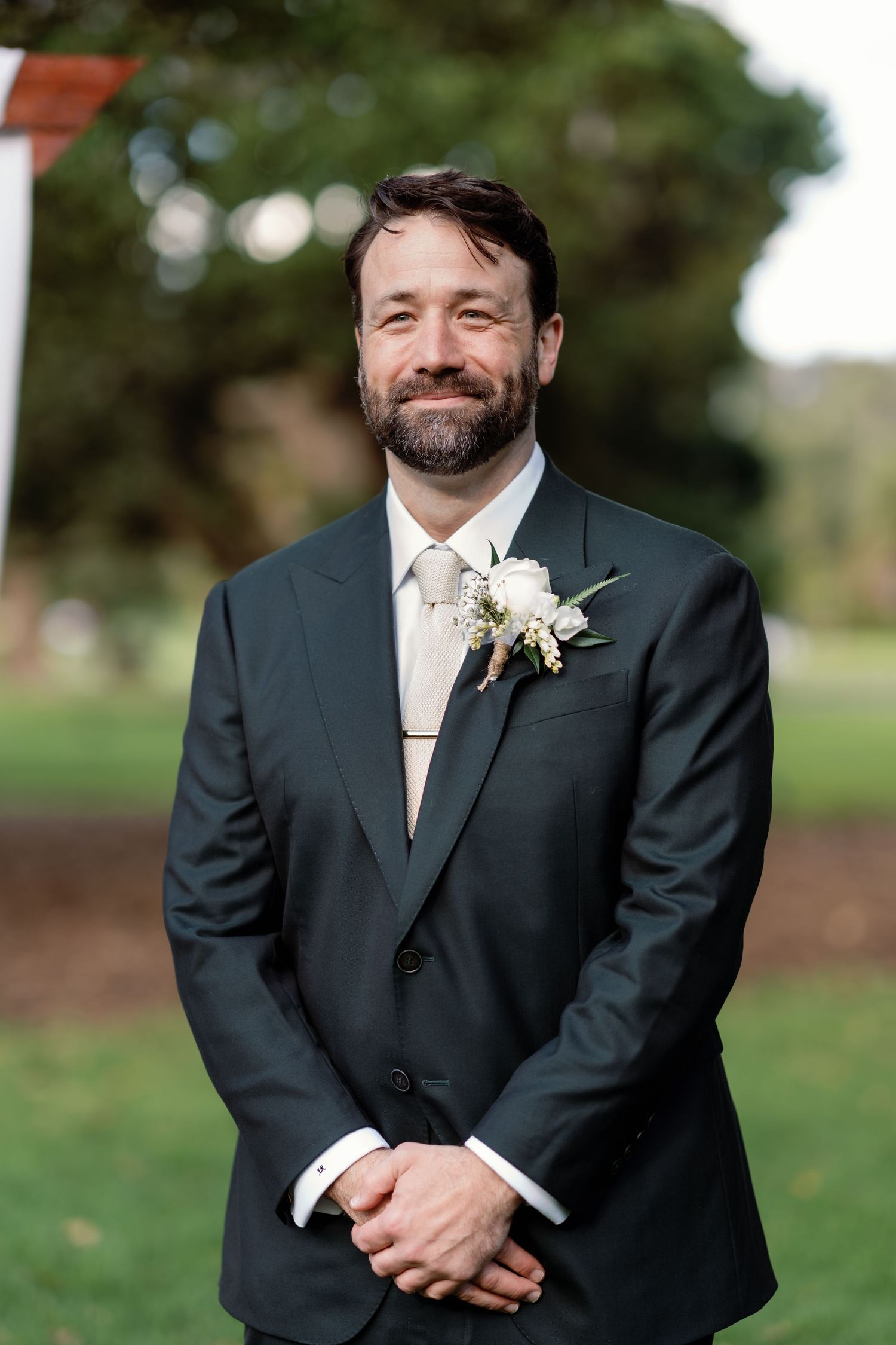 Smiling groom in dark suit with white boutonniere and gray tie poses outdoors at wedding ceremony.