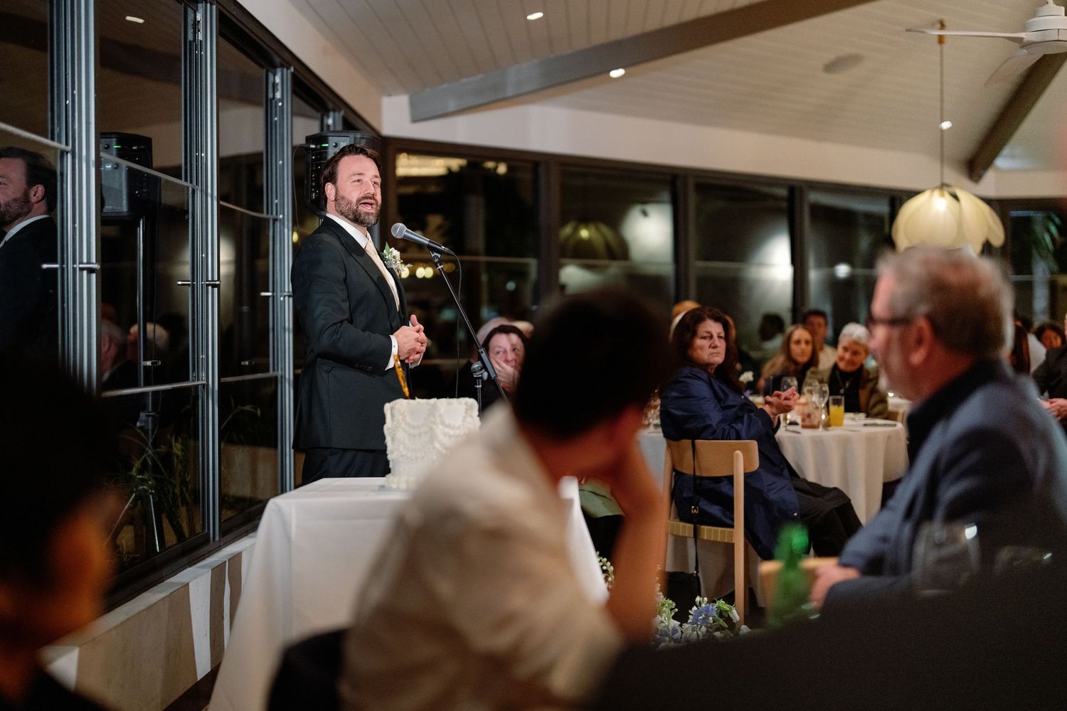 Man in suit giving speech at elegant wedding reception dinner with guests seated at tables.