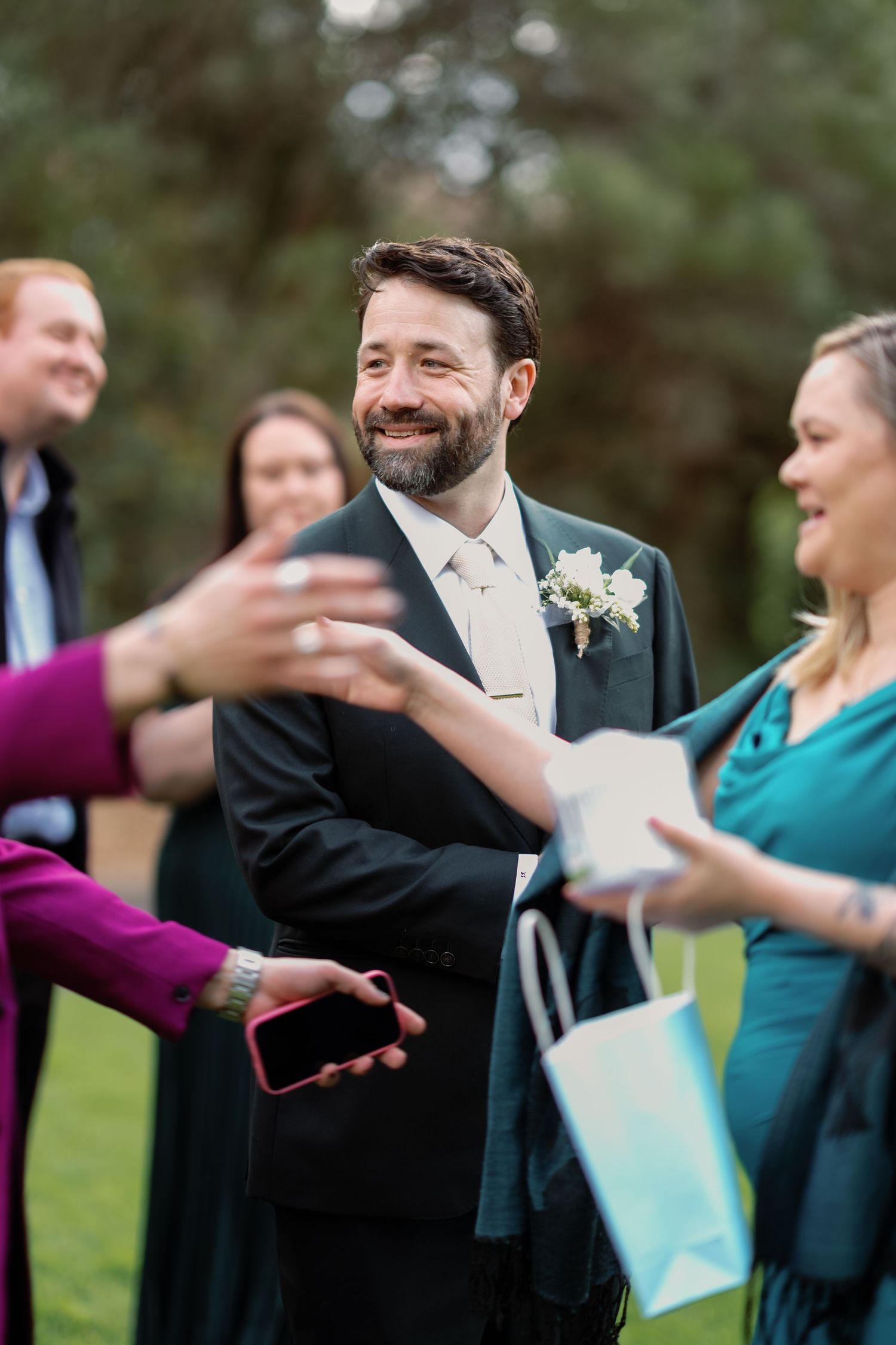 Smiling groom in dark suit with boutonniere receiving gifts from guests at an outdoor wedding celebration.
