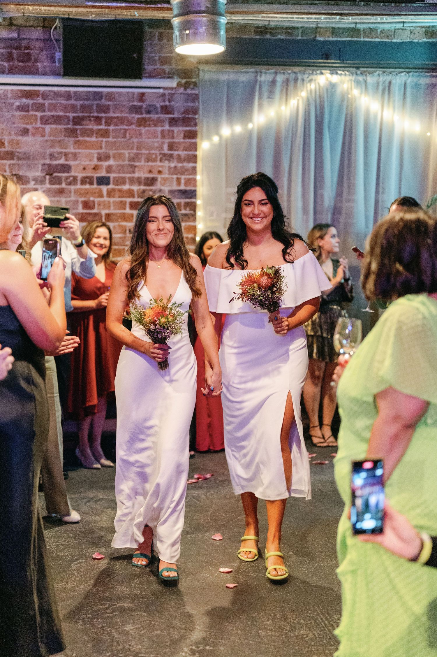Wedding party in white dresses walking and dancing at an industrial brick venue with string lights.