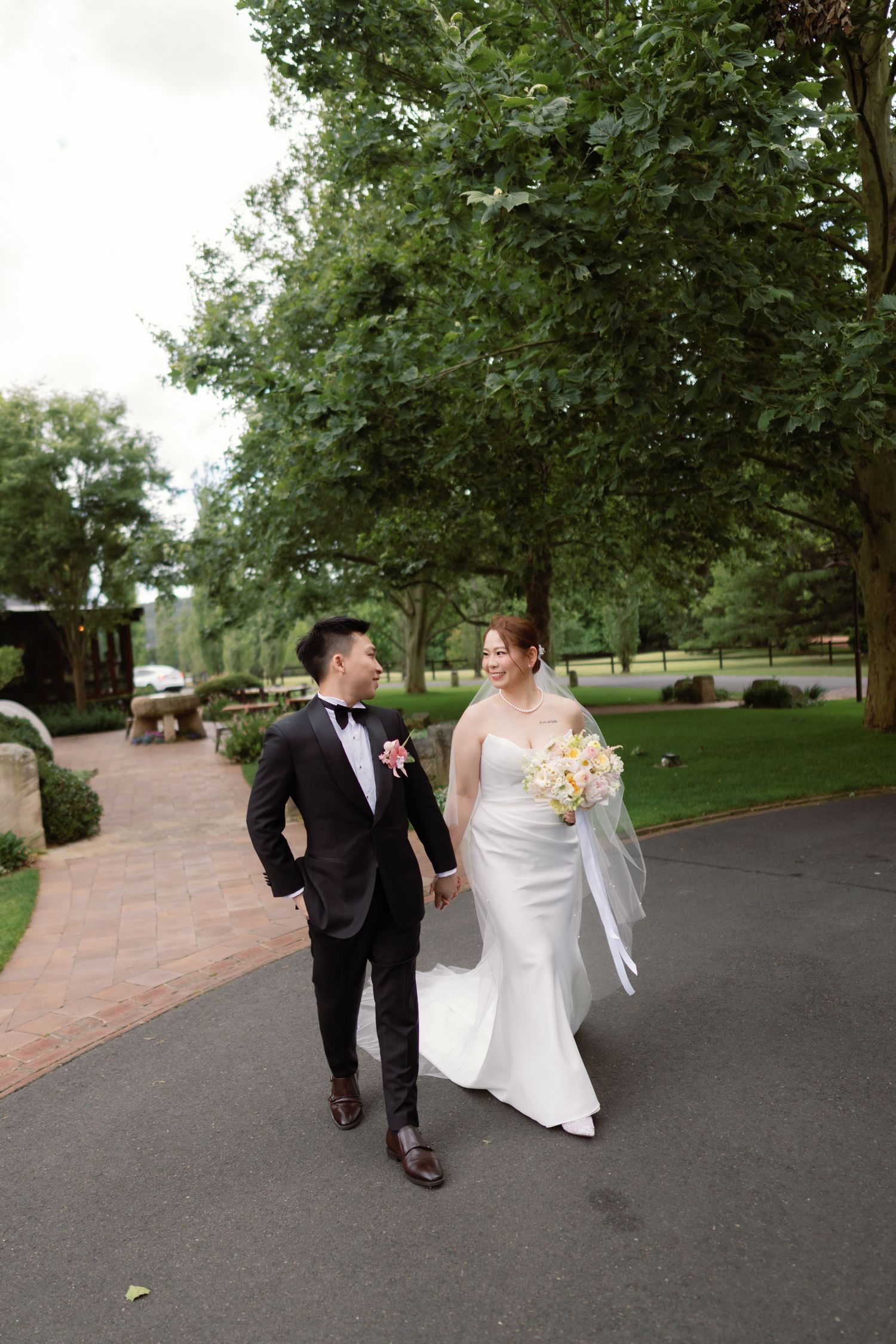 Couple strolls down paved pathway surrounded by lush green trees and landscaping.