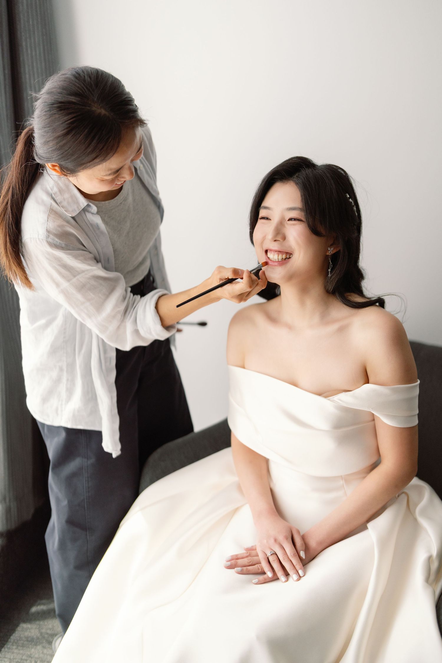 Bride in white off-shoulder gown getting ready with help from stylist in intimate indoor setting.