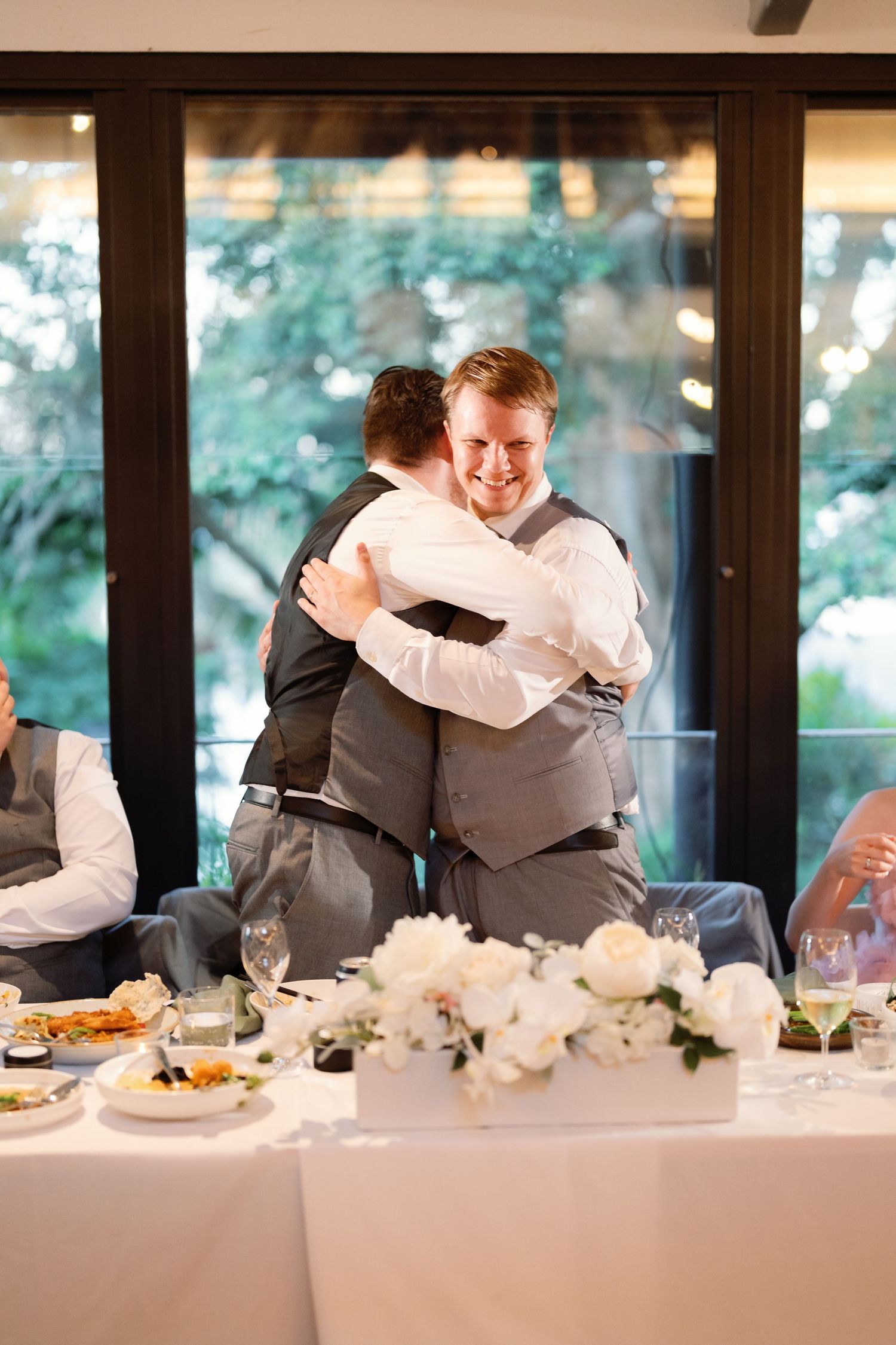 Wedding reception table setting with white flowers and guests sharing emotional moment at head table.